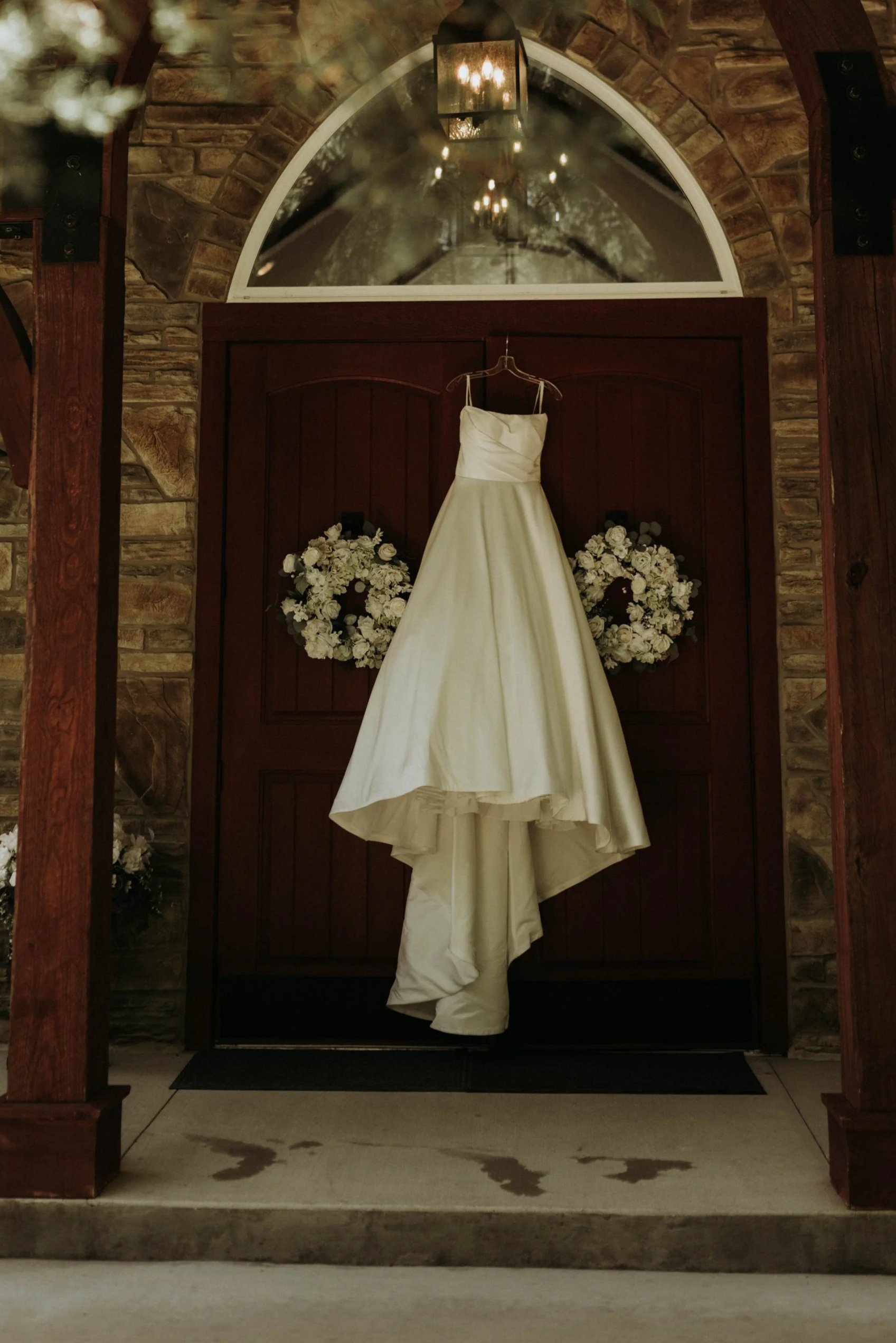 A wedding dress hanging on a hanger in front of a wooden door with floral arrangements on each side. The scene is inside a building with stone walls and a large, arched window above the door. A chandelier with candles is reflected in the window.
