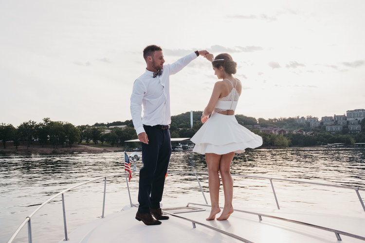 A couple on a boat, with the man twirling the woman in a white dress, by a river during sunset.