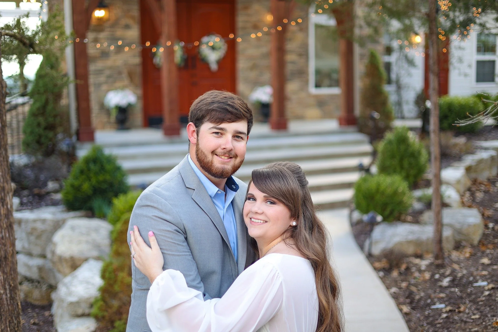 A young couple dressed in formal attire, smiling and embracing outdoors in front of a house with string lights and decorated porch.