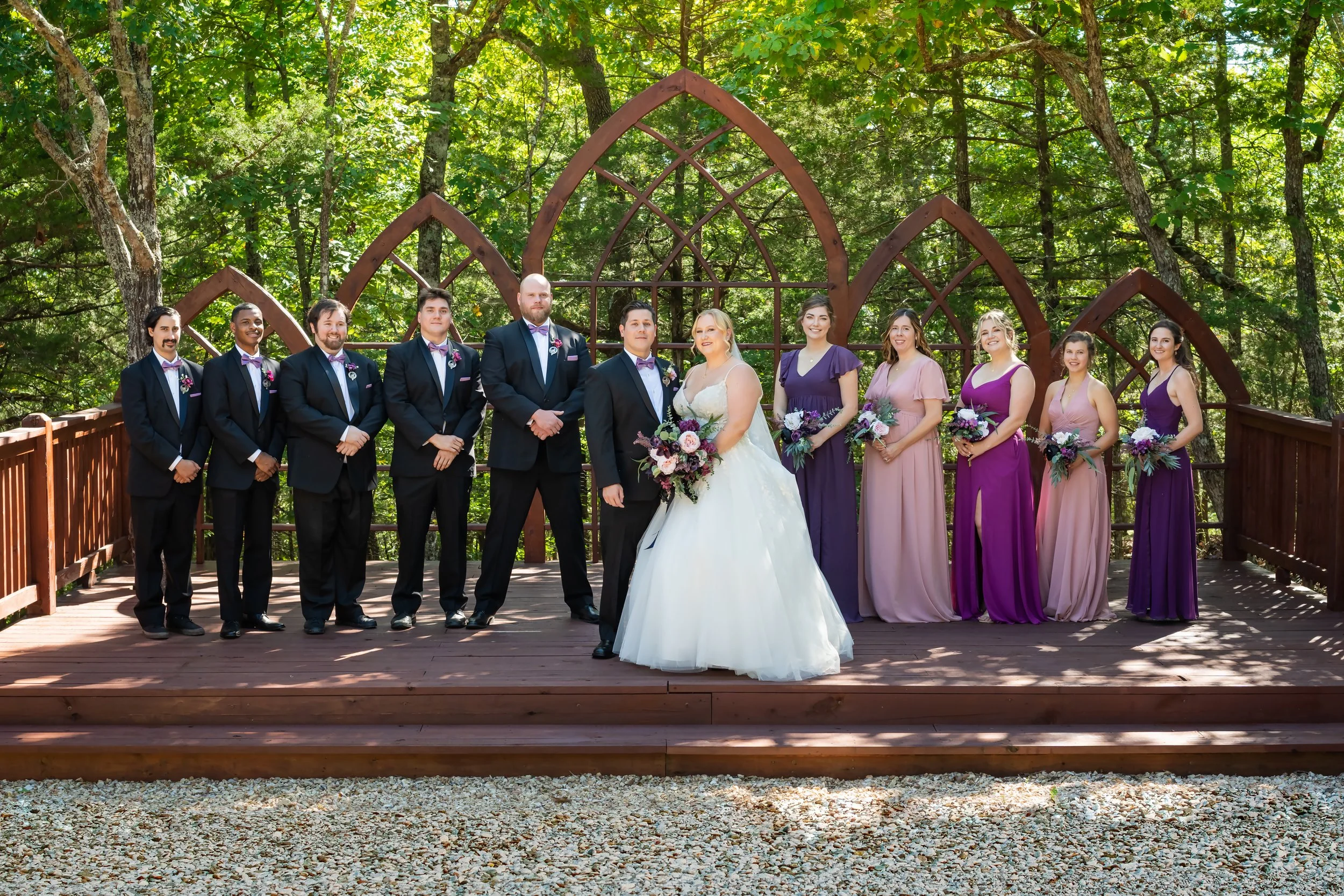 A wedding party stands on a wooden platform in a forest, with the bride and groom in the center, surrounded by bridesmaids and groomsmen, all dressed in formal attire, with the women wearing purple and pink dresses and holding bouquets, and the men i