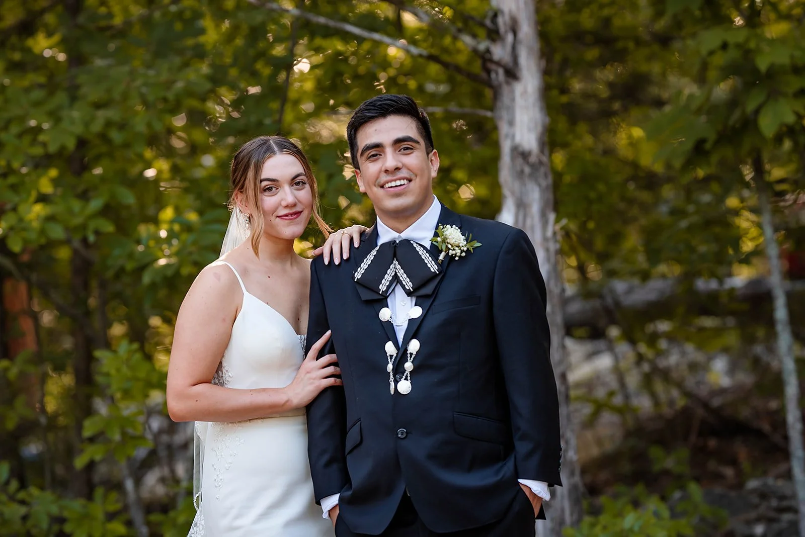 A bride and groom standing outdoors, smiling at the camera, surrounded by green trees, at their wedding.