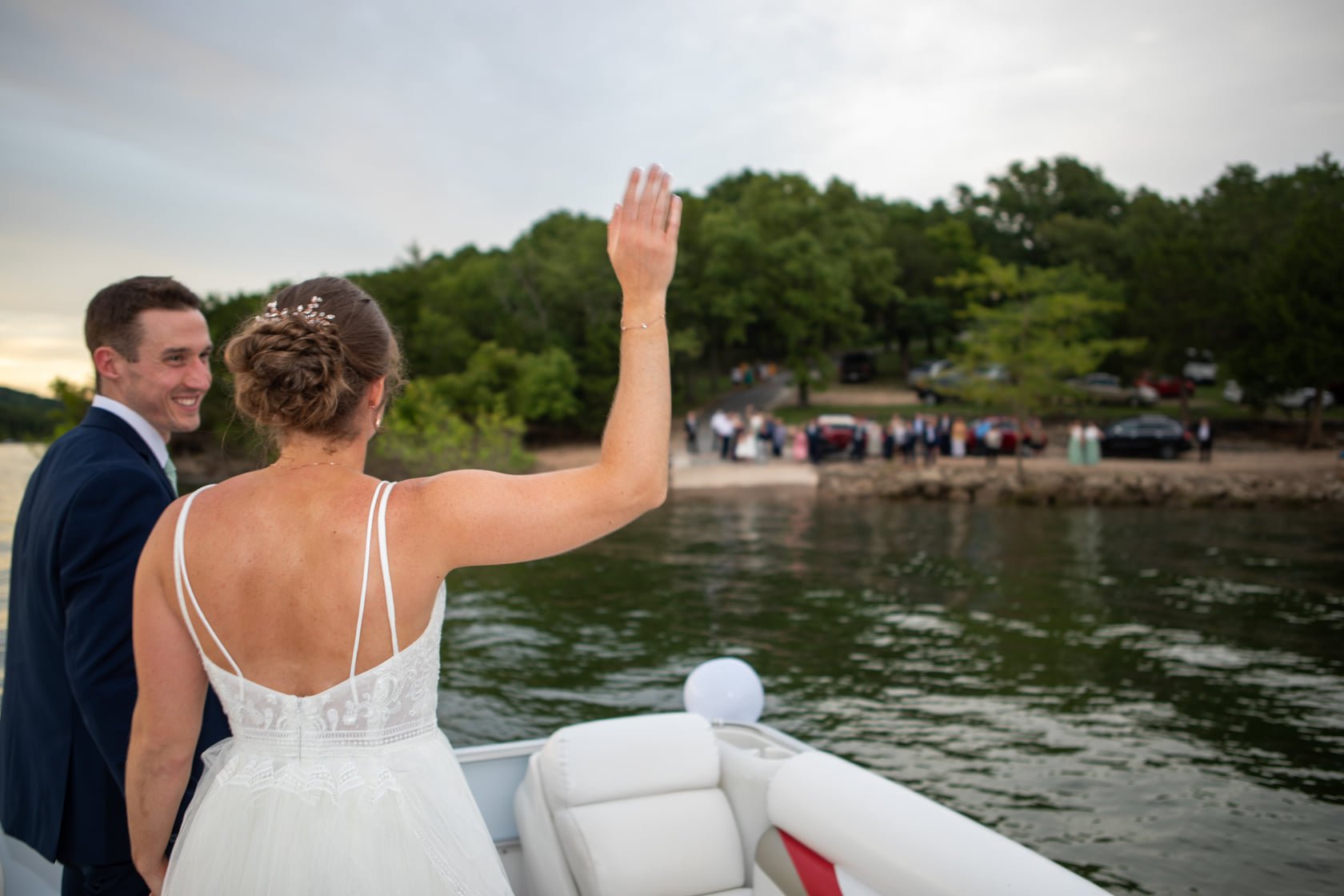 A bride and groom on a boat, waving goodbye, with a group of people on the shore in the background