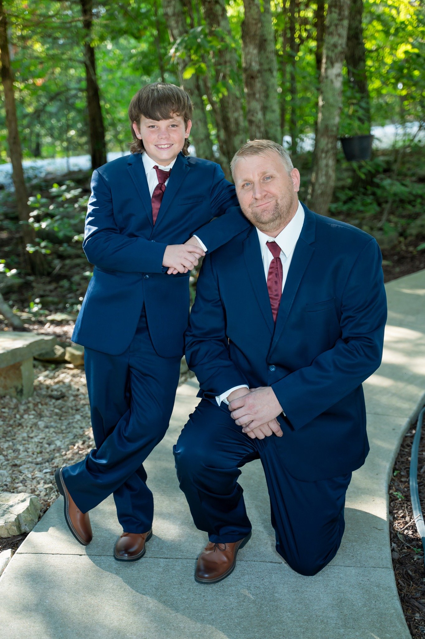 A man and a boy wearing blue suits, white shirts, and maroon ties posing outdoors in a wooded area. The boy is standing with one arm around the man's shoulder and the other hand on his hip, smiling. The man is kneeling on one knee with his hands clasped, looking at the camera with a slight smile.