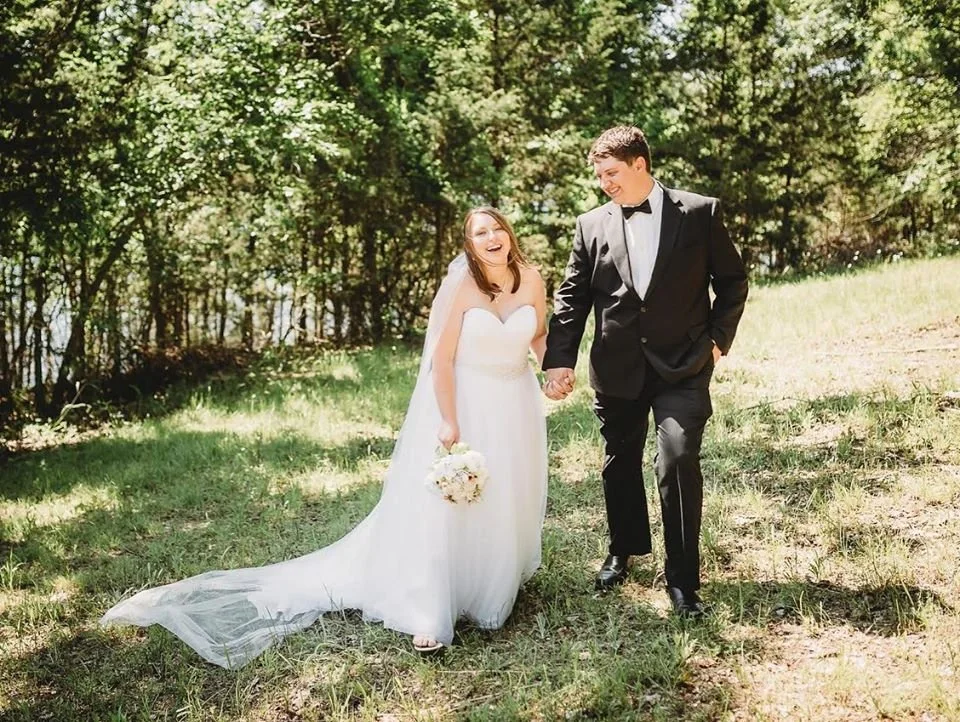 A bride and groom walking hand in hand outdoors in a wooded area, dressed in wedding attire, smiling and enjoying the moment.