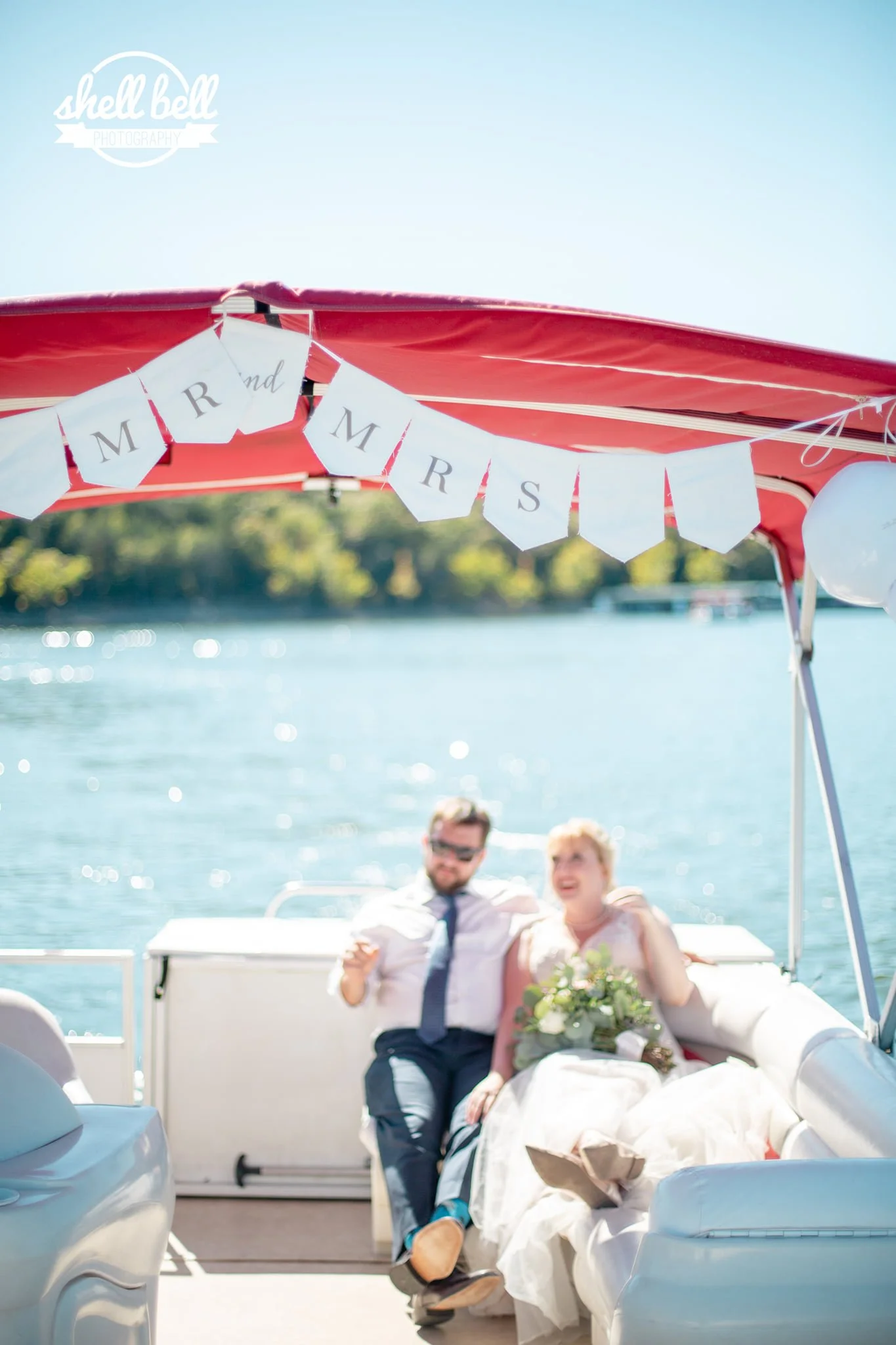 A couple dressed in wedding attire, sitting on a boat with water and trees in the background, celebrating their wedding with a "Mrs." and "Mrs." bunting hanging overhead.