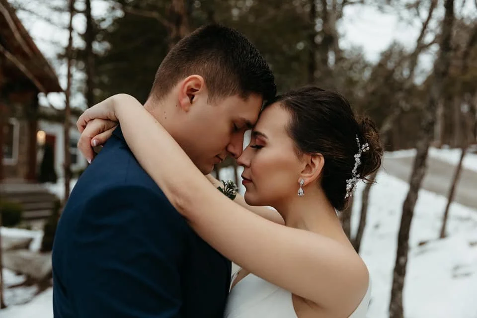 A newlywed couple embraces outdoors during winter, with both their eyes closed, standing close with foreheads touching, in a snow-covered landscape with trees and a house in the background.