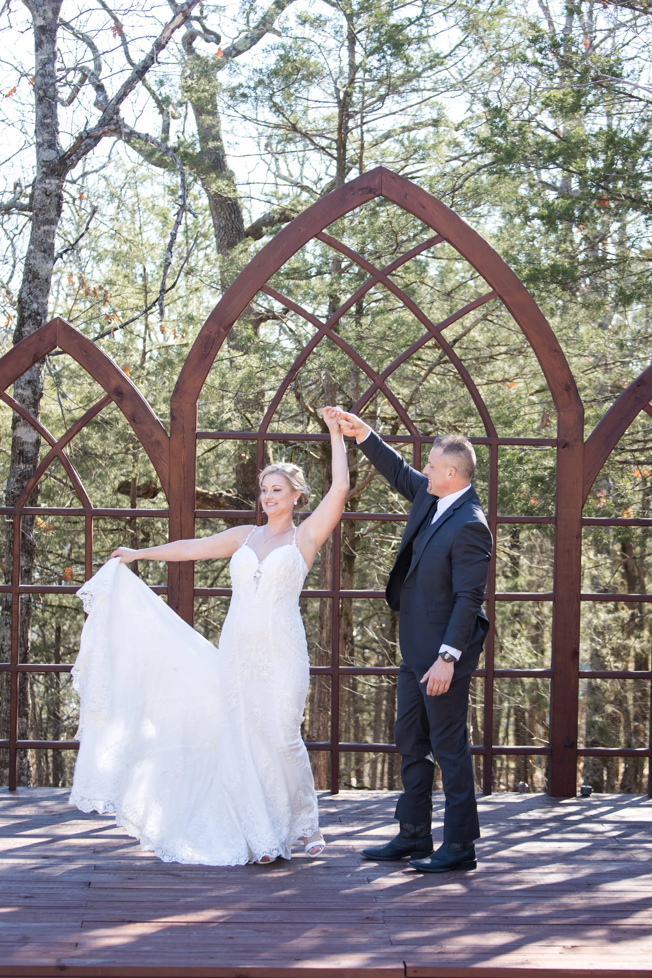 A bride and groom dancing outdoors on a wooden stage, with trees in the background. The bride is wearing a white wedding dress and the groom is in a black suit. They are holding hands, smiling, and appear happy.