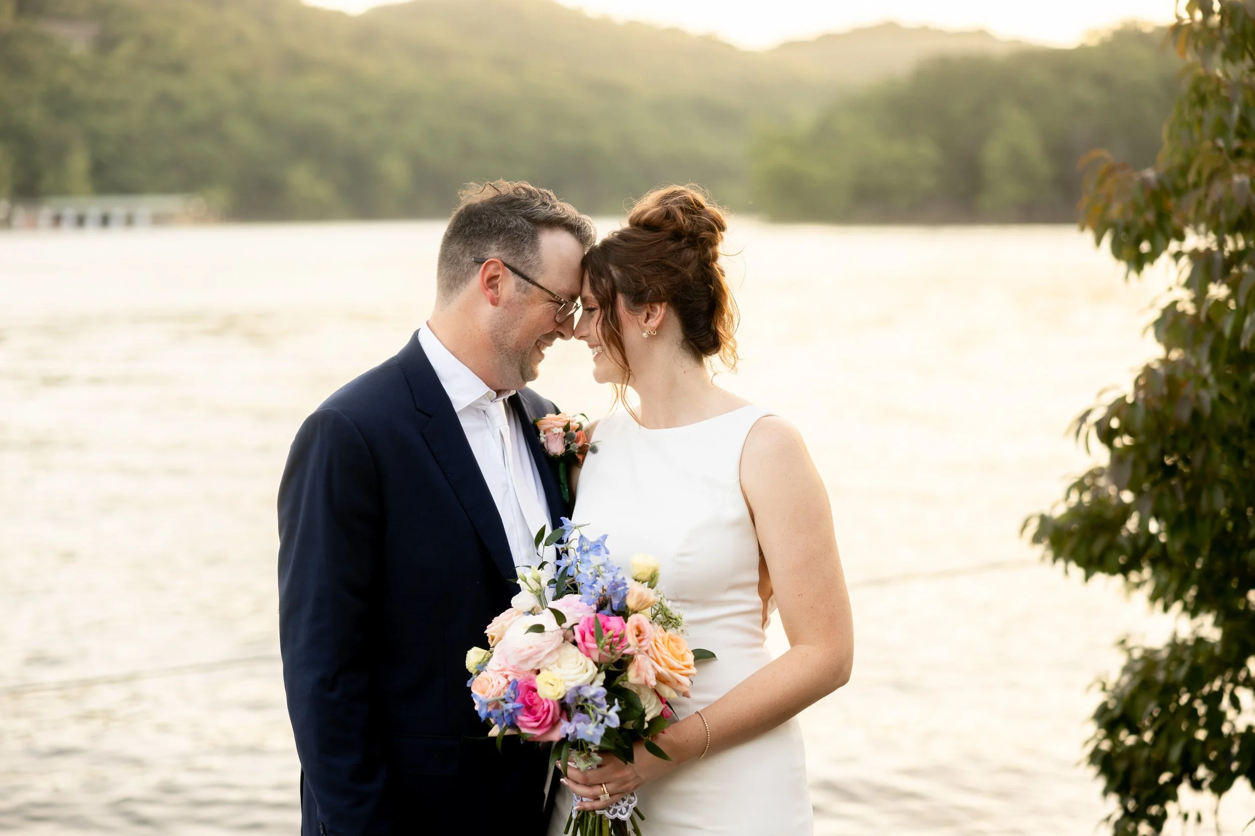 A newlywed couple, a man in a dark suit and glasses and a woman in a white dress, sharing a kiss by a body of water at sunset, with trees in the background and a branch with leaves on the right.