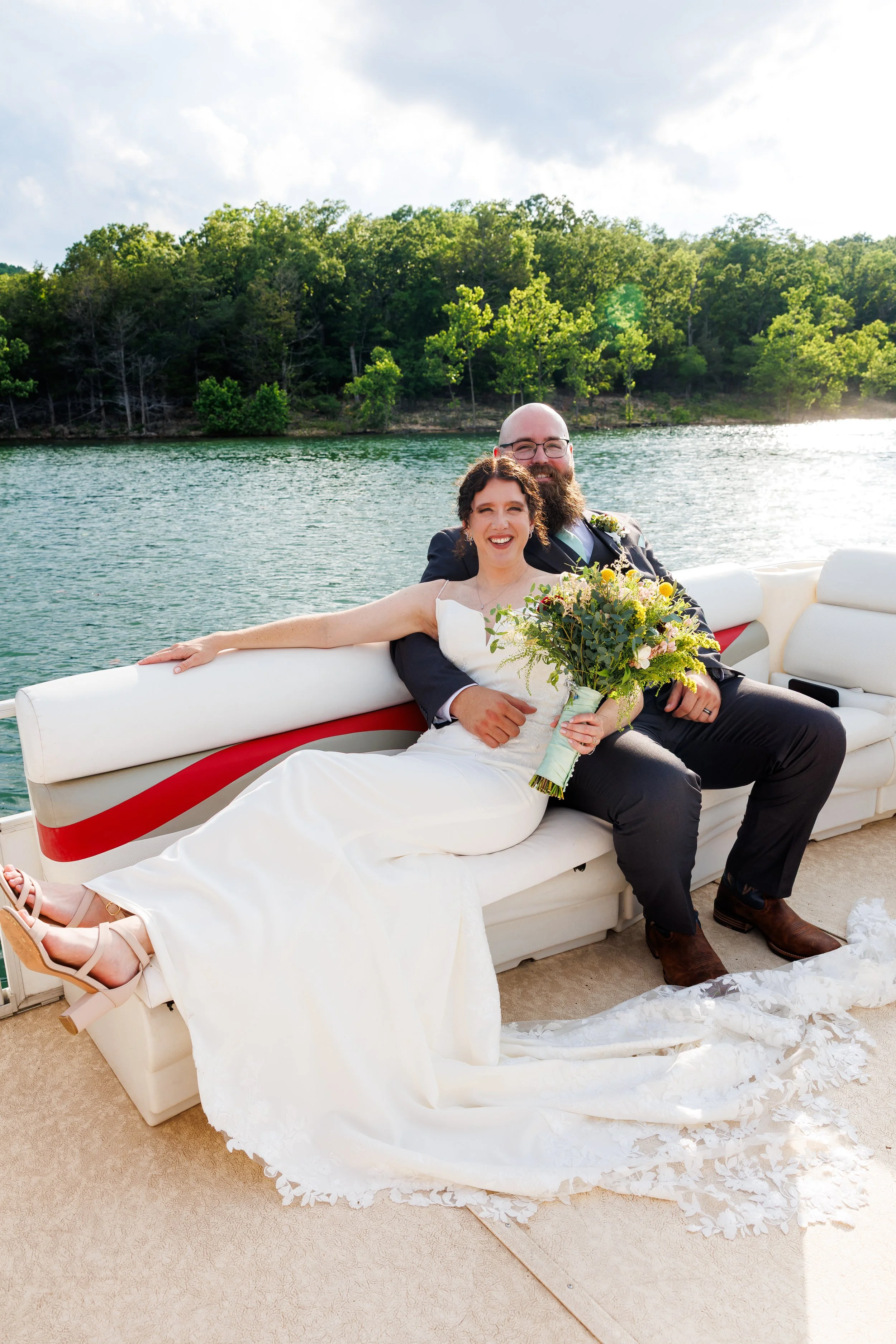 A bride and groom on a boat ride celebrating their wedding, with the bride holding a bouquet and the groom wearing glasses and a suit, in front of a wooded shoreline with water and cloudy sky in the background.