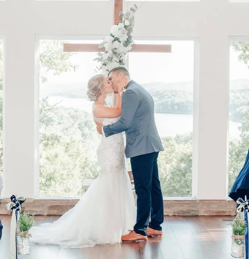 A bride and groom share a kiss during their wedding ceremony inside a building with large windows and a scenic view of water and trees in the background.