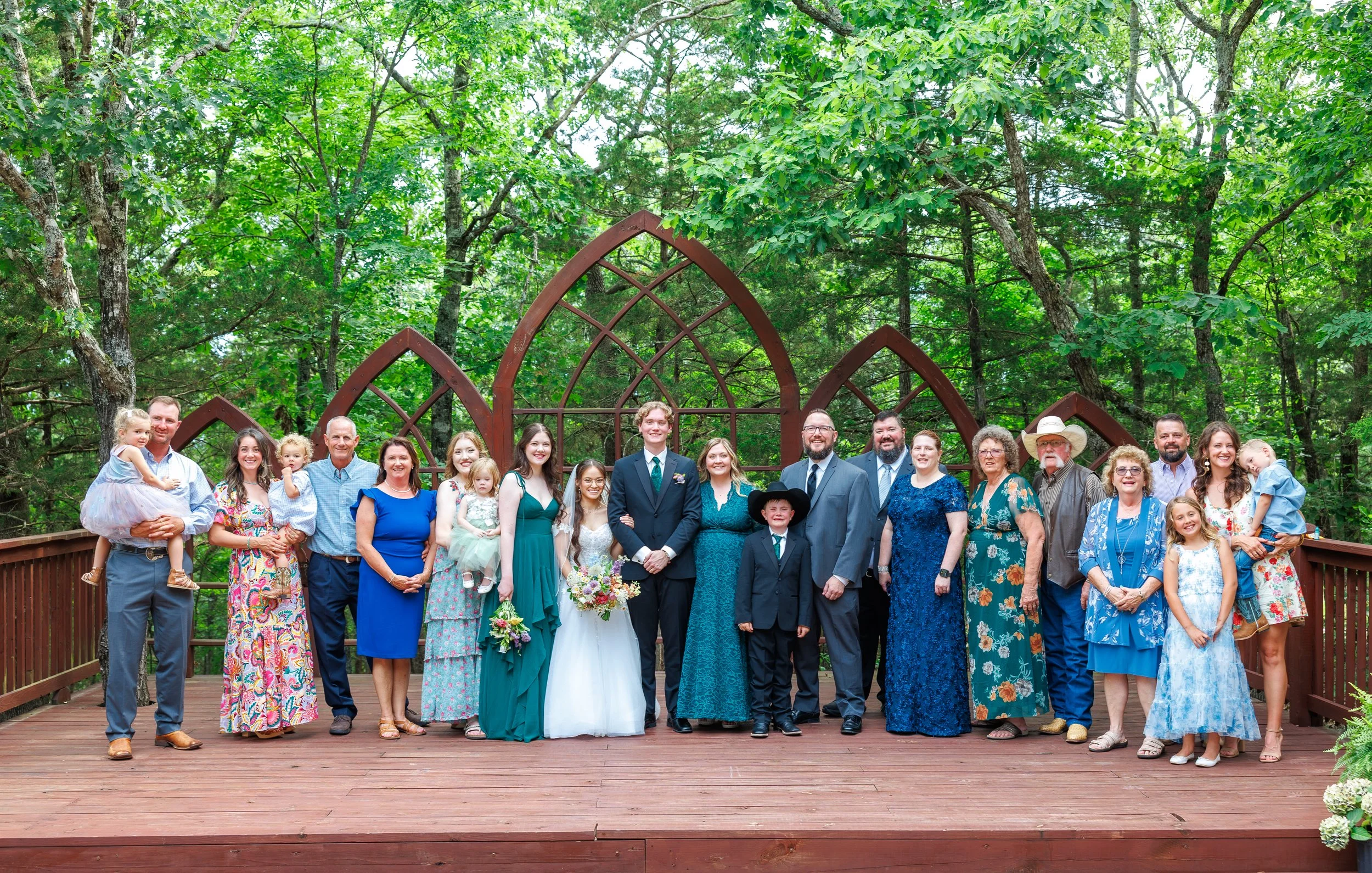 A large group of people, including children, adults, and seniors, pose for a photo on a wooden deck surrounded by green trees. The group is dressed in colorful, semi-formal attire, suggesting a celebratory occasion such as a wedding.