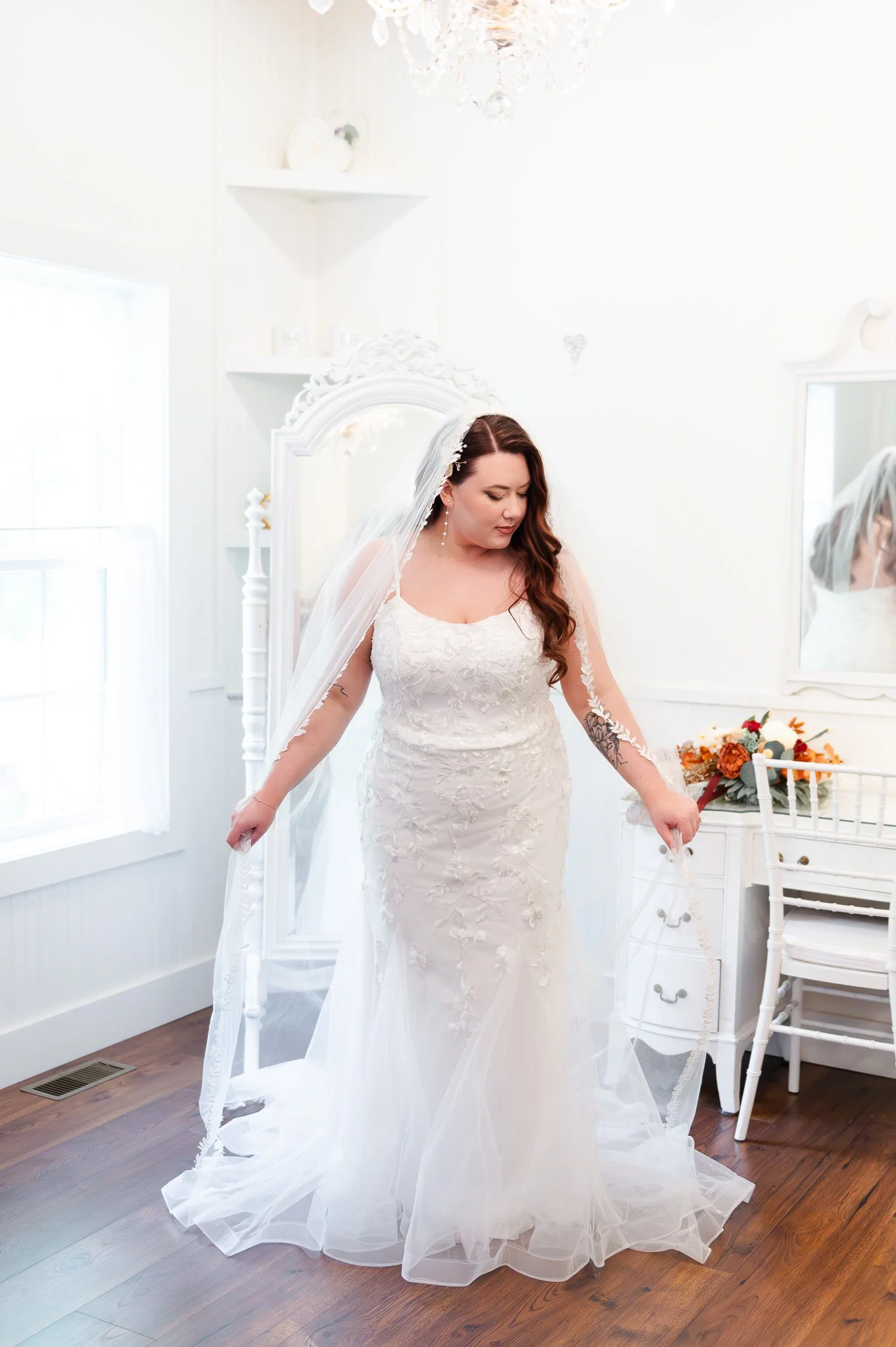A bride with long brown hair in a white lace wedding dress, looking down at her dress, in a bright, white room with a chandelier and a white dresser with flowers.