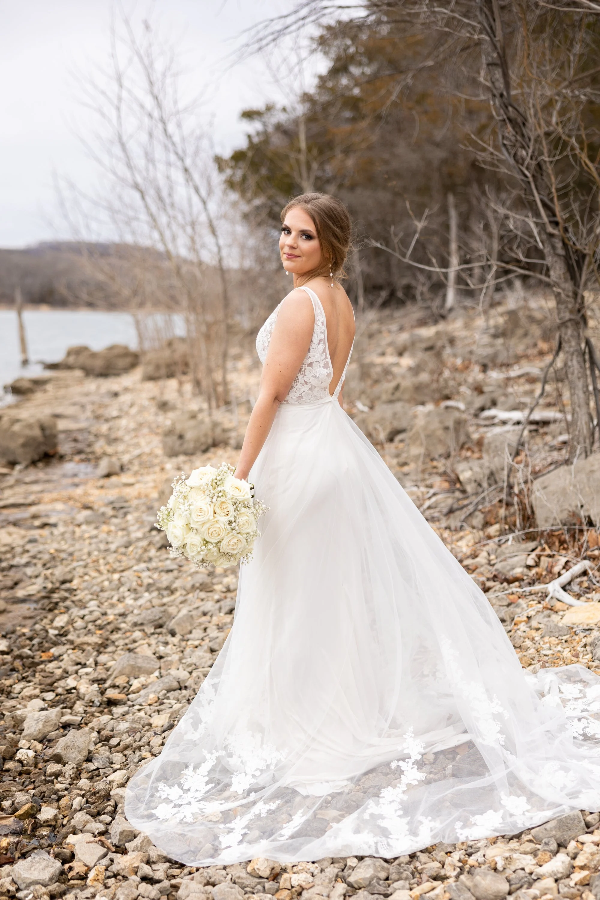 A woman in a white wedding dress holding a bouquet of white roses, standing on rocky terrain near a body of water with leafless trees in the background.