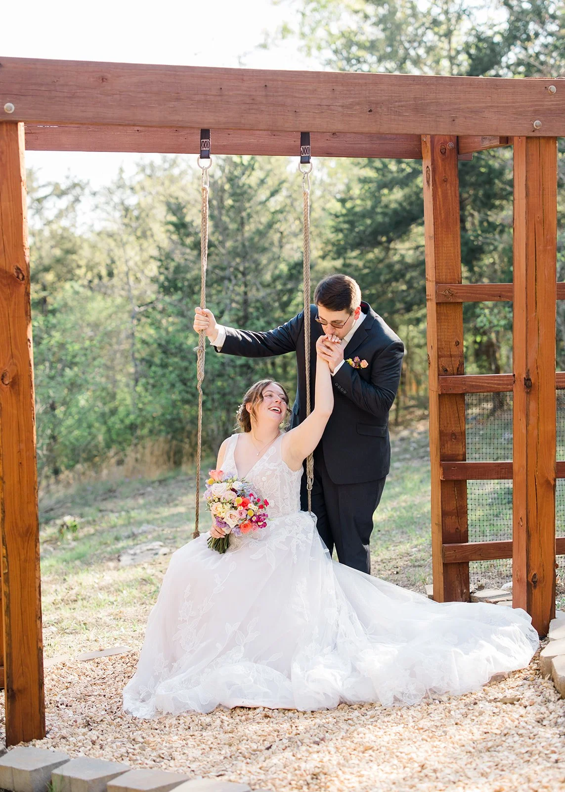 Bride sitting on a swing holding a bouquet of flowers, smiling at groom. Groom standing beside her, gently kissing her hand. Outdoor setting with trees in the background, during daytime.