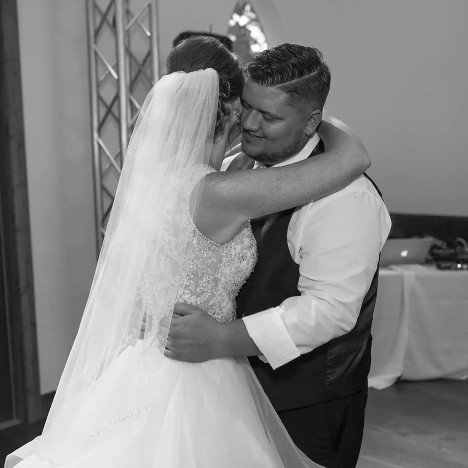 A bride and groom embrace during their wedding, with the bride wearing a lace wedding dress and veil, and the groom in a white shirt and dark vest, in a room with a DJ table in the background.