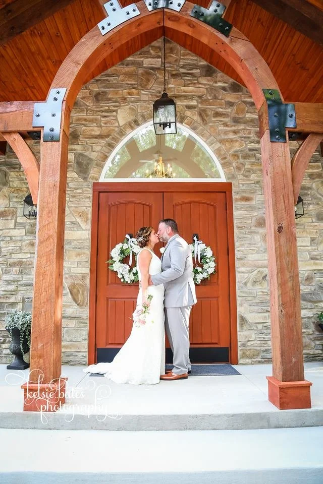 A bride and groom standing close together, about to kiss, in front of a large wooden door decorated with white floral wreaths, inside a building with a stone wall and wooden archway.