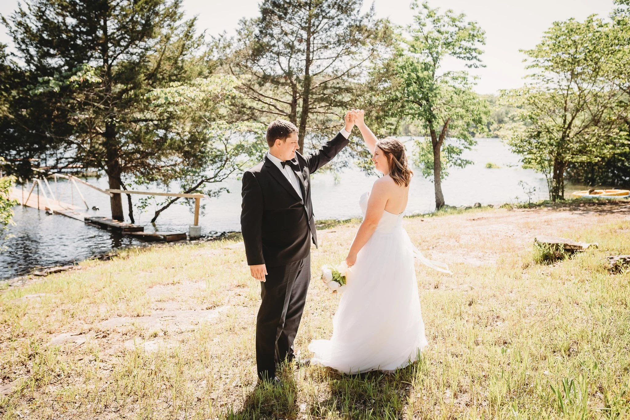 A bride and groom dancing outdoors near a lake with trees, the groom in a black tuxedo and the bride in a white wedding dress holding a bouquet.