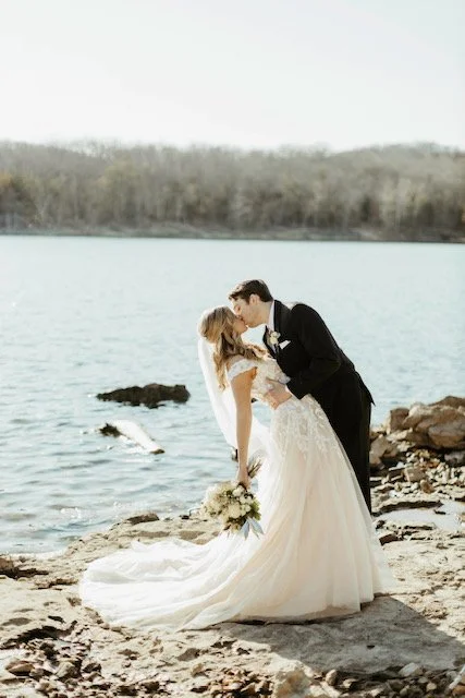 A bride and groom Kiss on a rocky shoreline by the water, with the bride holding a bouquet and the groom in a tuxedo.