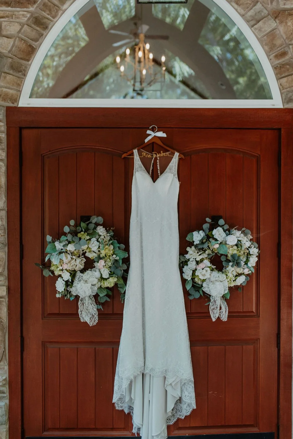 A white wedding dress hanging on a wooden door, flanked by two floral wreaths with white and pale pink flowers and green leaves, with a chandelier and arched window reflected in the glass above.