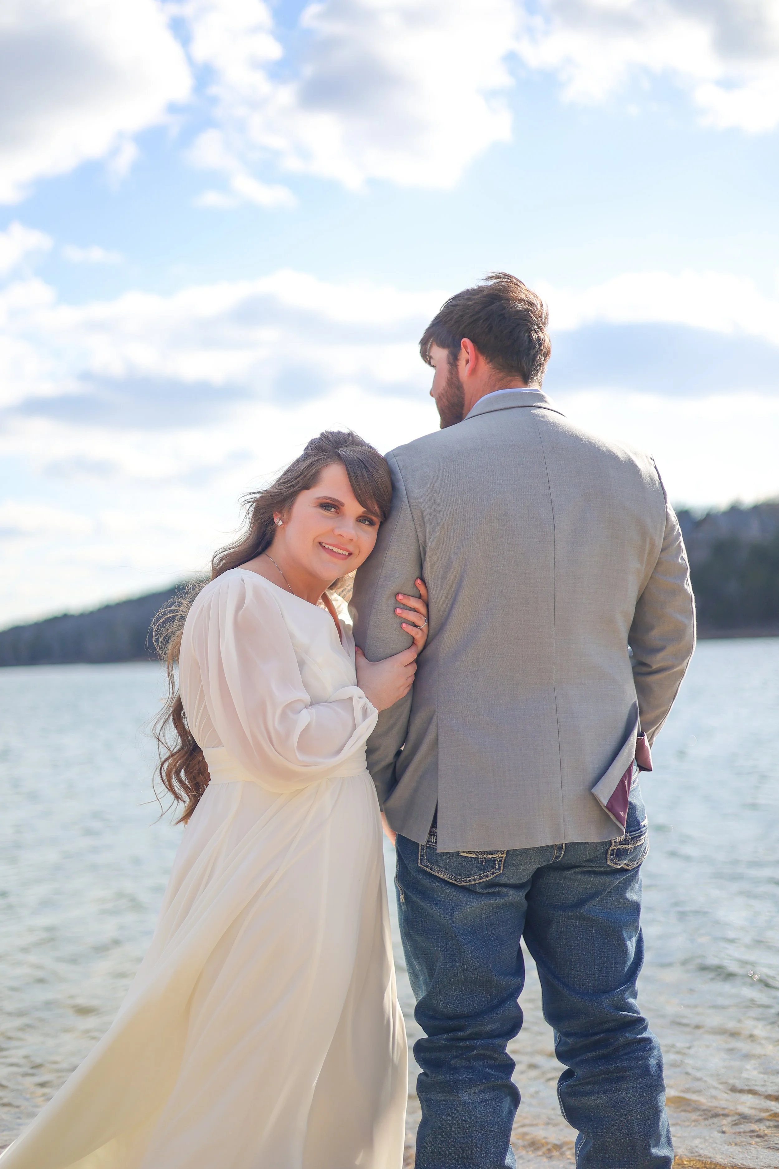 A smiling woman in a white dress holding hands with a man in a gray blazer and jeans, standing by a lake with hills and a partly cloudy sky in the background.
