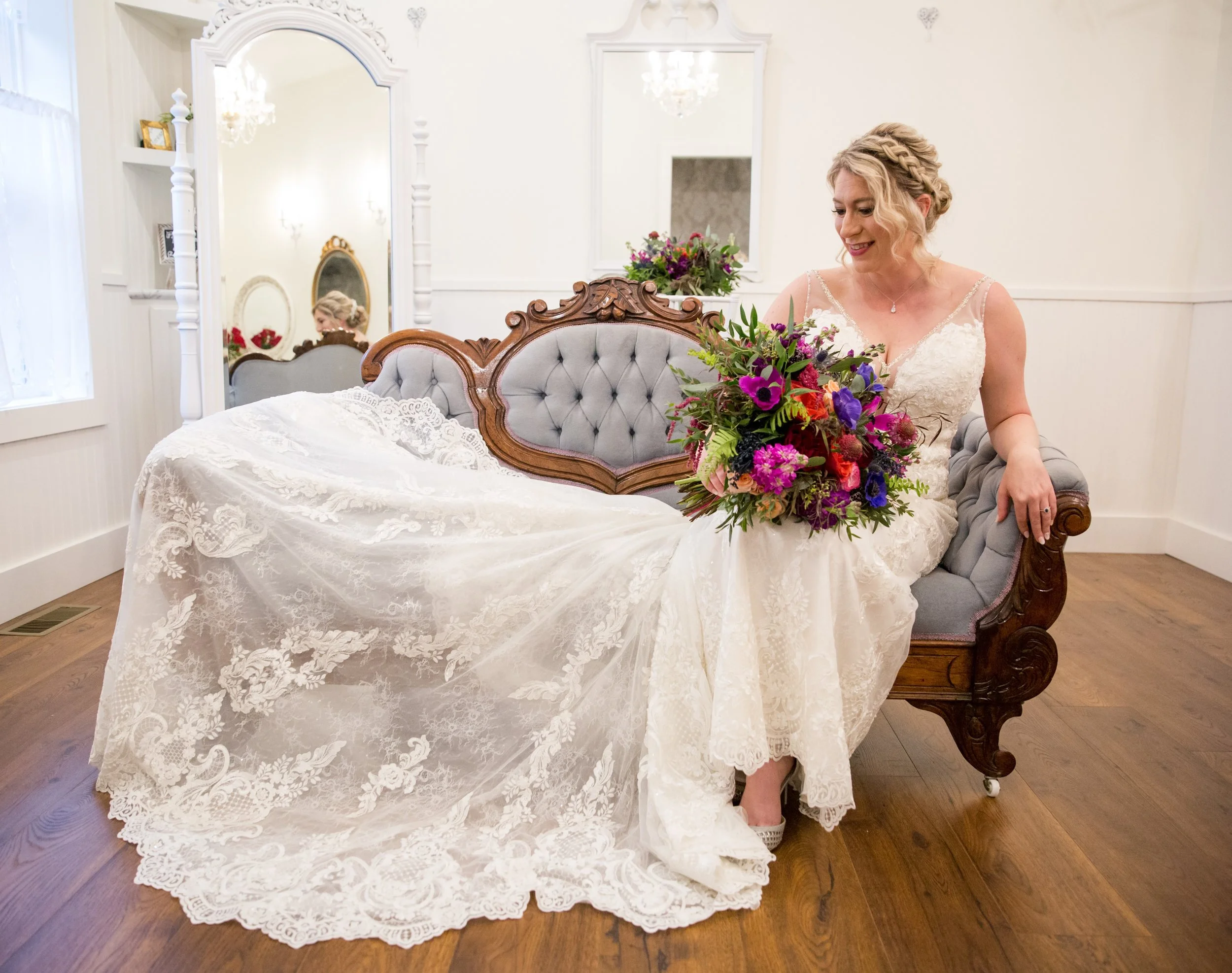 A bride sitting on a vintage gray upholstered and carved wooden bed, holding a colorful flower bouquet, indoors with white walls and mirrors.