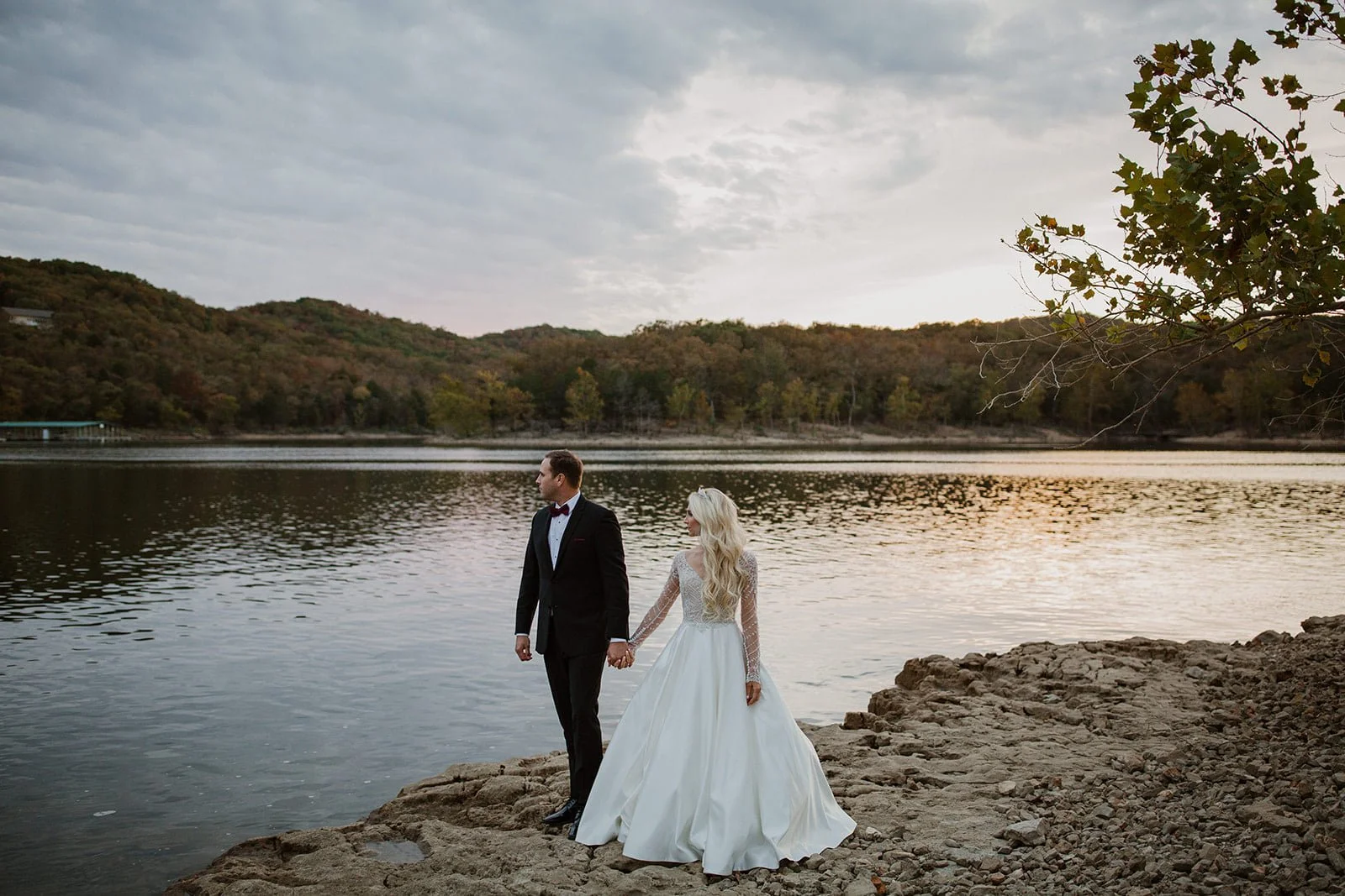 A bride and groom holding hands on a rocky lakeside at sunset, with hills and trees in the background.