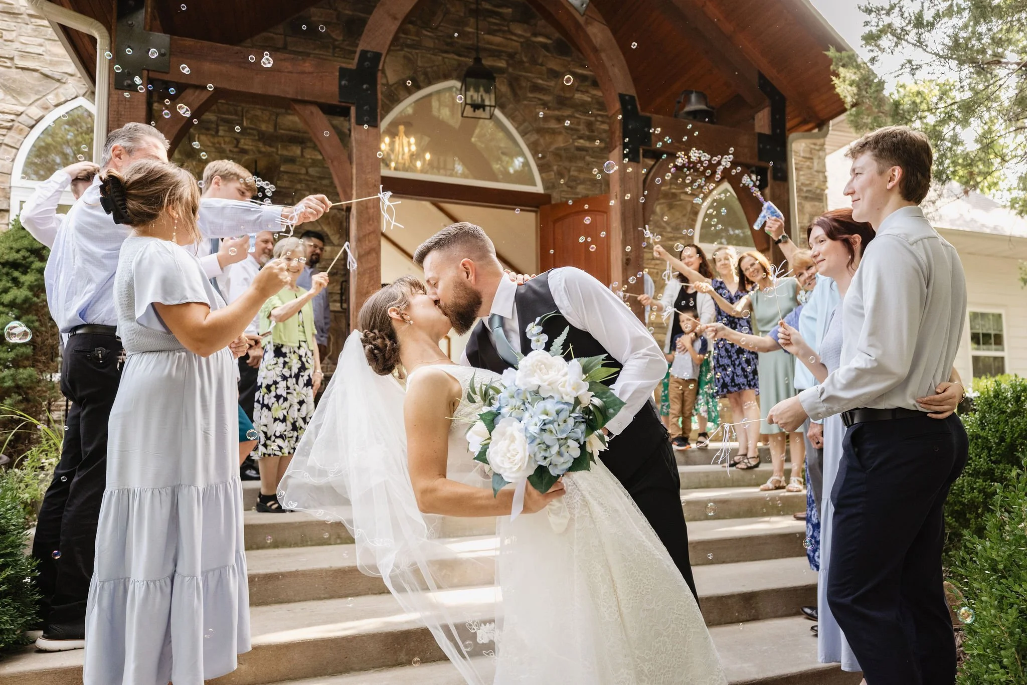 A couple kisses on the steps outside a stone house while surrounded by guests celebrating with bubbles during a wedding.