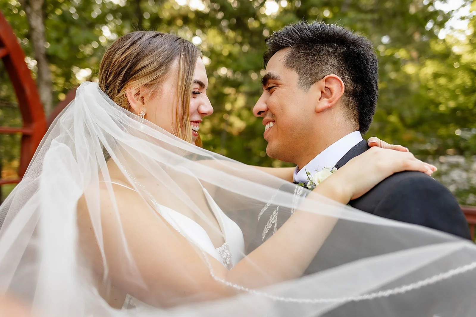 A bride and groom smiling, facing each other, with the bride's veil draped around both of them, against a blurred background of green trees.