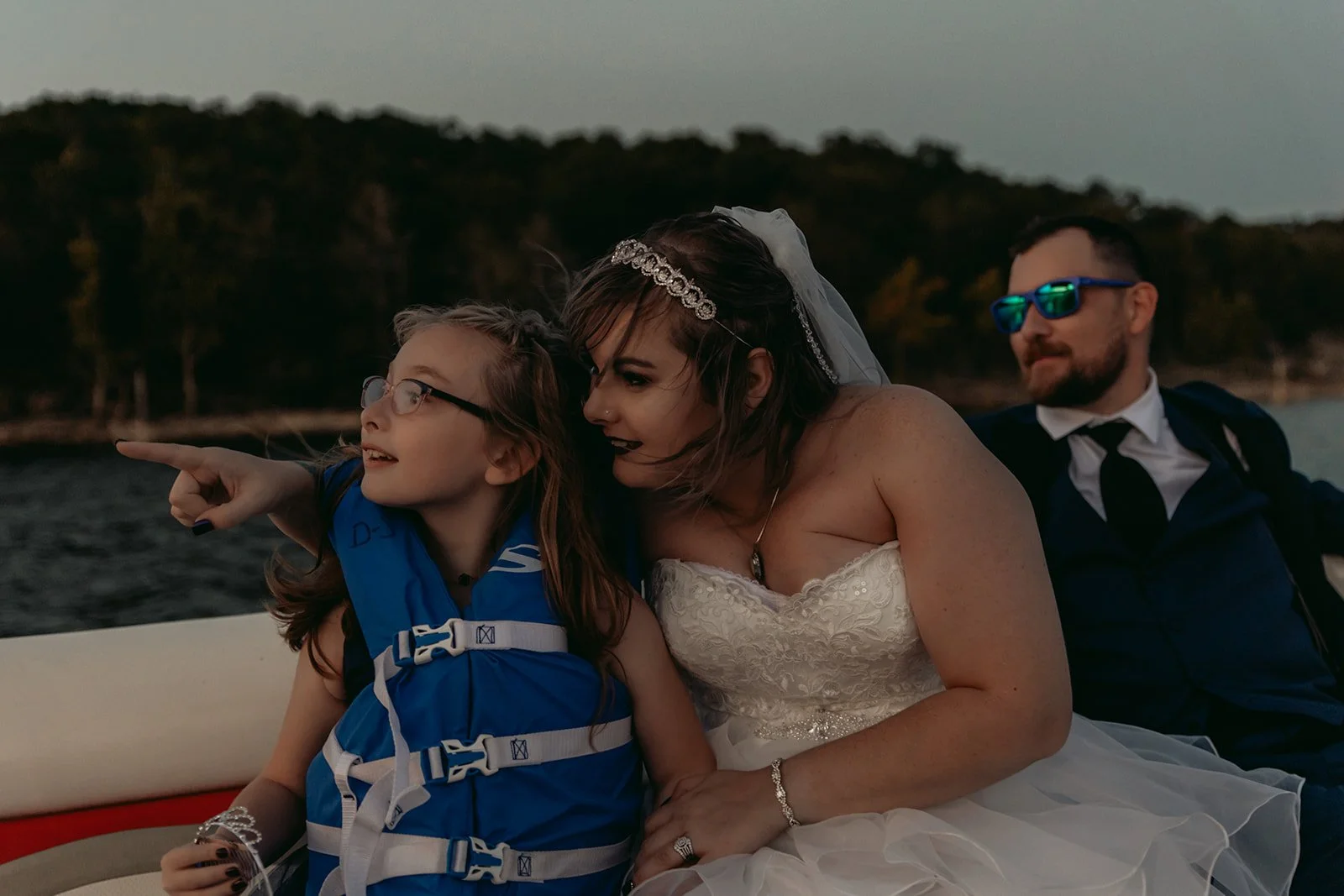 A woman in a wedding dress and a veil, with a necklace and bracelet, is sitting beside a young girl wearing glasses and a blue life jacket, on a boat at sunset. The woman and girl are looking in the same direction, and the woman is pointing towards s