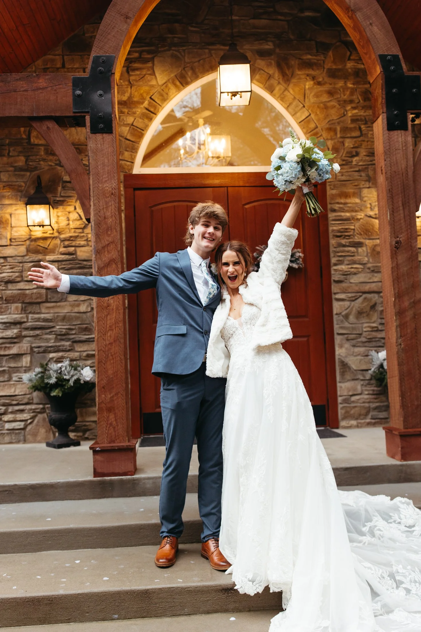 A happy couple in wedding attire stands outside a wooden and stone building, celebrating. The woman is holding a bouquet and wearing a white wedding gown and a fur jacket. The man is wearing a blue suit and brown shoes. Both are smiling, with the wom