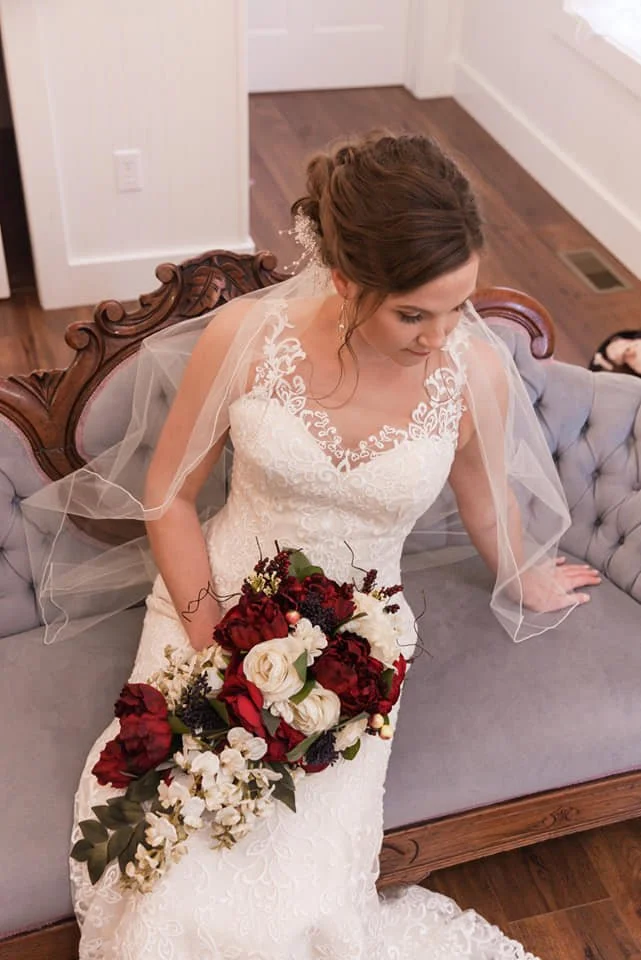 Bridal bride in lace wedding dress holding a bouquet of red and white flowers sitting on a vintage gray and wood sofa.