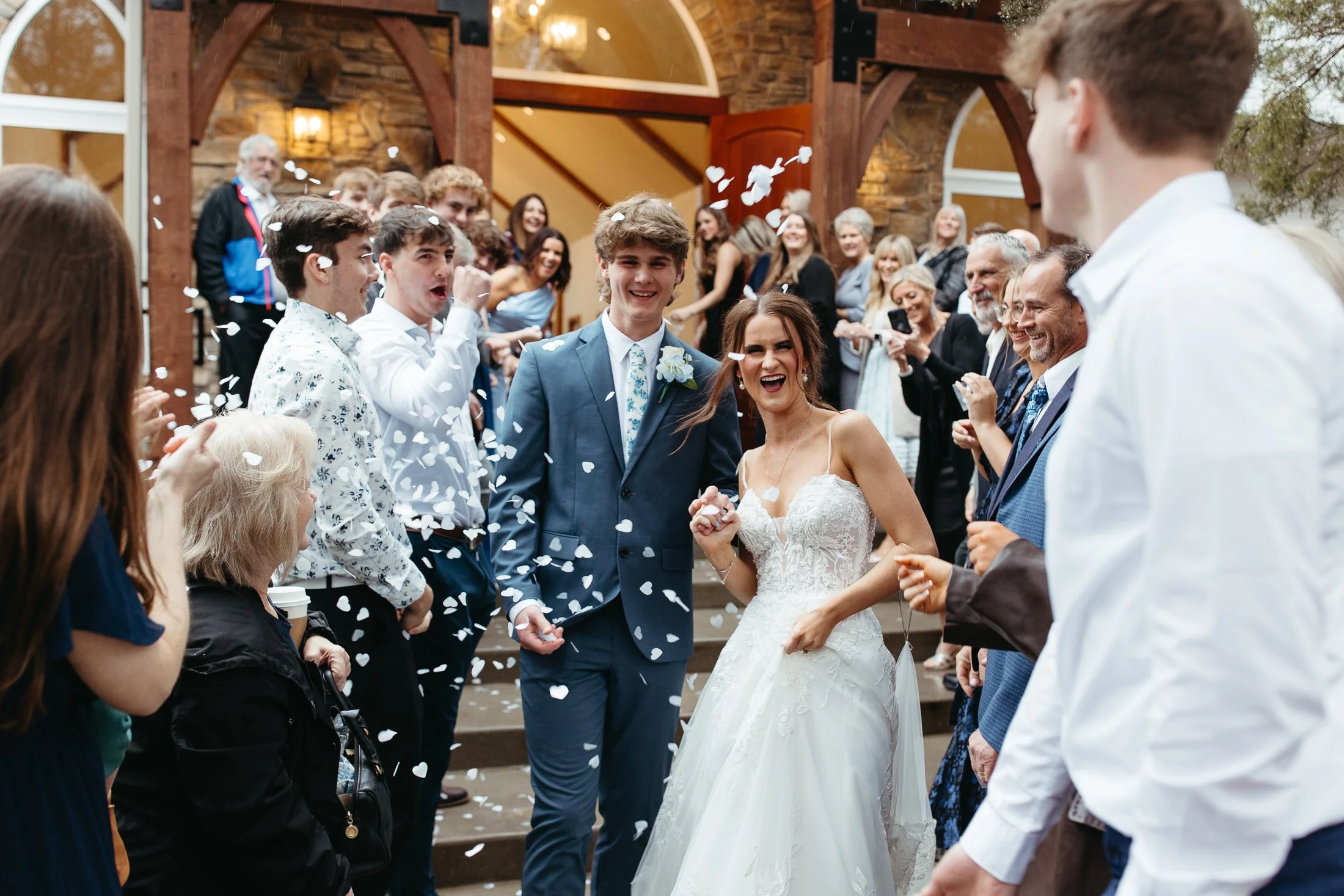 A bride and groom smiling and holding hands while walking through a crowd of guests throwing white heart-shaped confetti. The scene appears to be at an outdoor wedding reception, with a stone and wood building in the background and guests smiling and