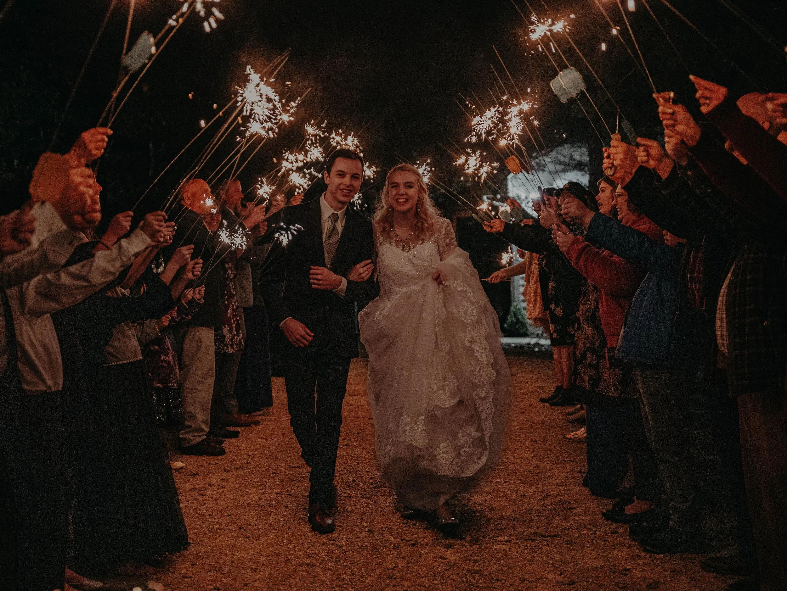 A bride and groom walking together at night, surrounded by a crowd holding sparklers during a celebration.