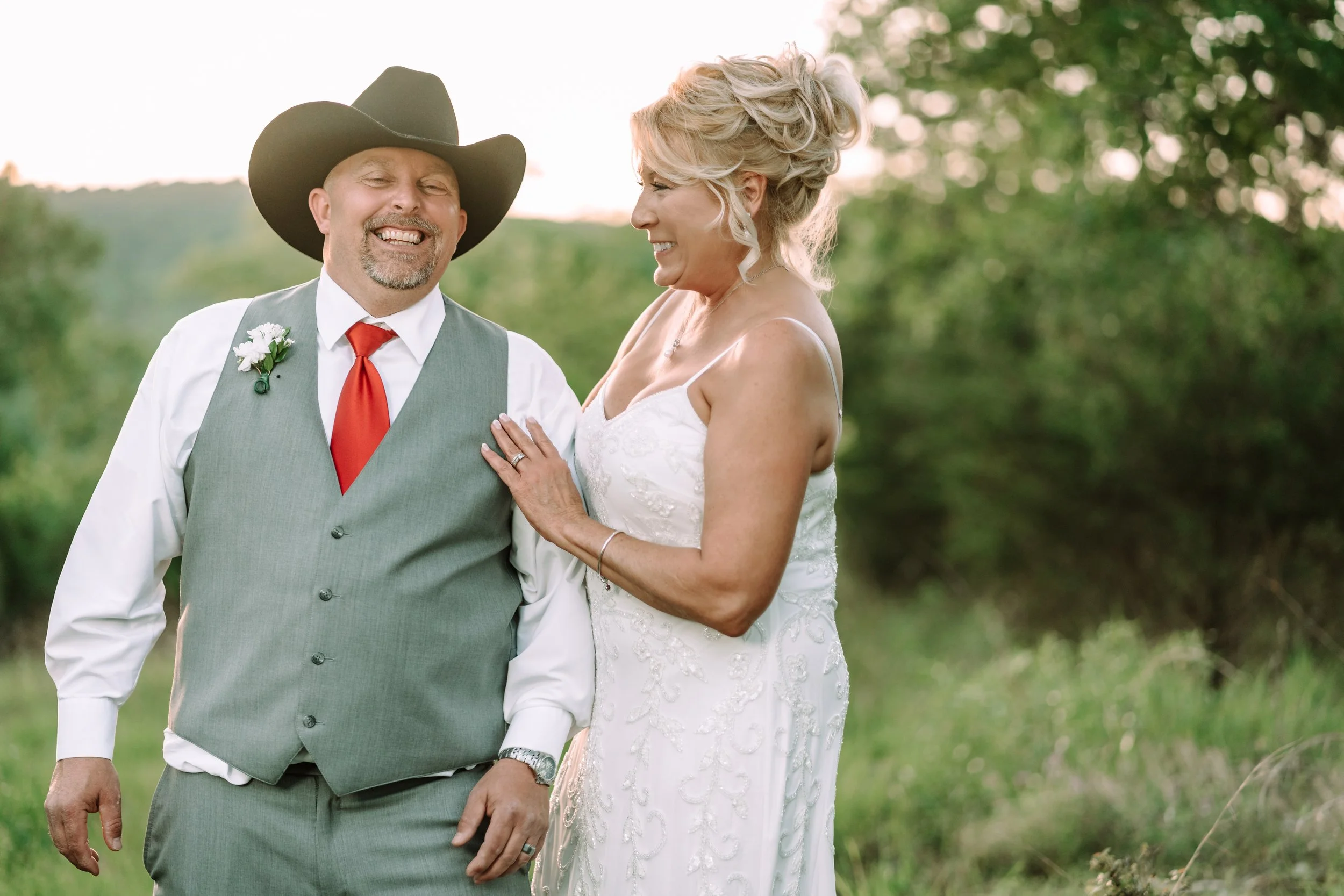 A smiling man wearing a cowboy hat, white shirt, red tie, gray vest, and boutonniere, standing outdoors with a woman in a white wedding dress, both looking at each other and smiling, with greenery and trees in the background during sunset.