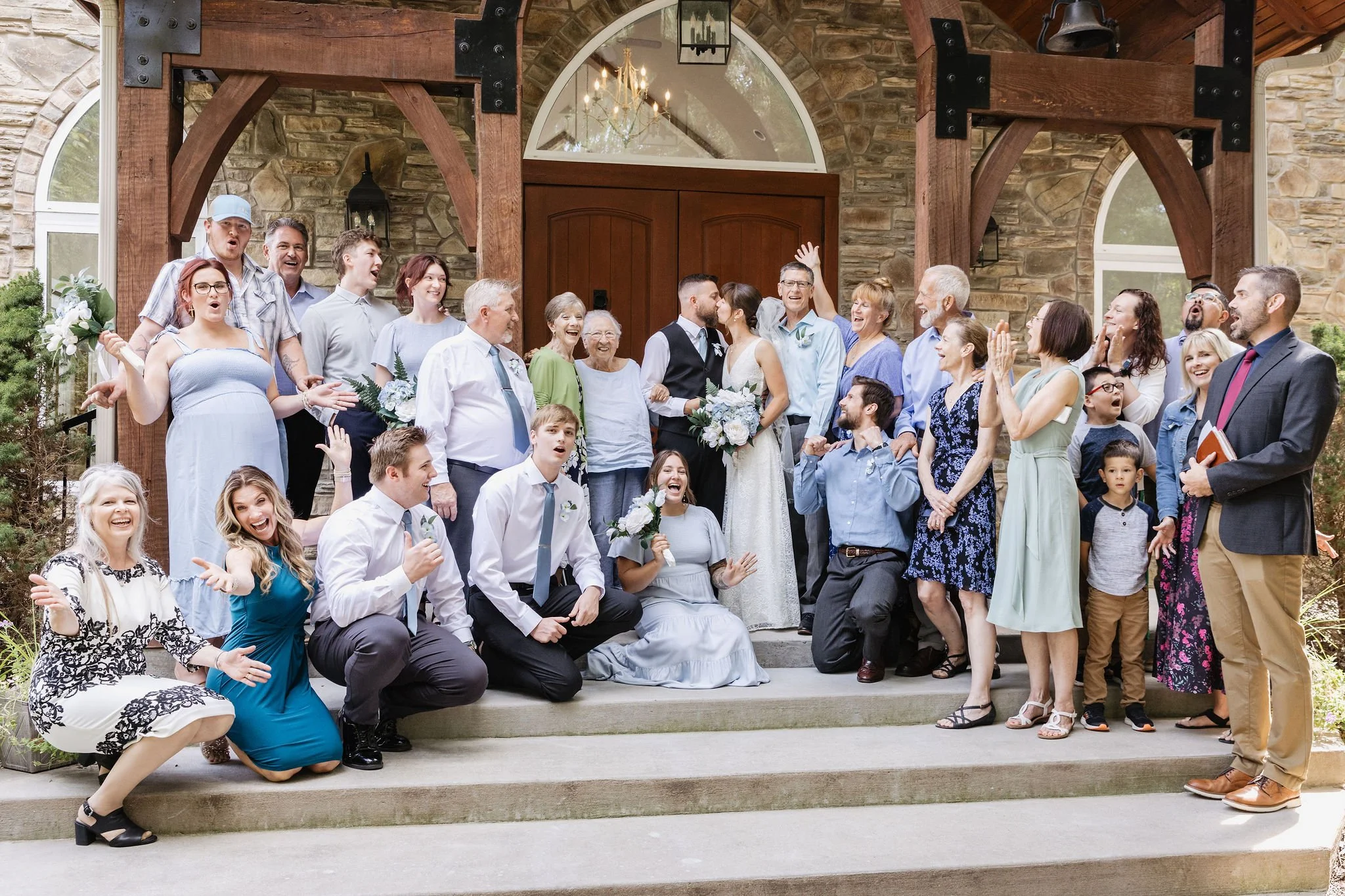 A wedding celebration with a large group of people gathered on a staircase outside a rustic stone building. The bride and groom are in the center, kissing, surrounded by smiling guests of various ages. Some guests are cheering, clapping, and making celebratory gestures.