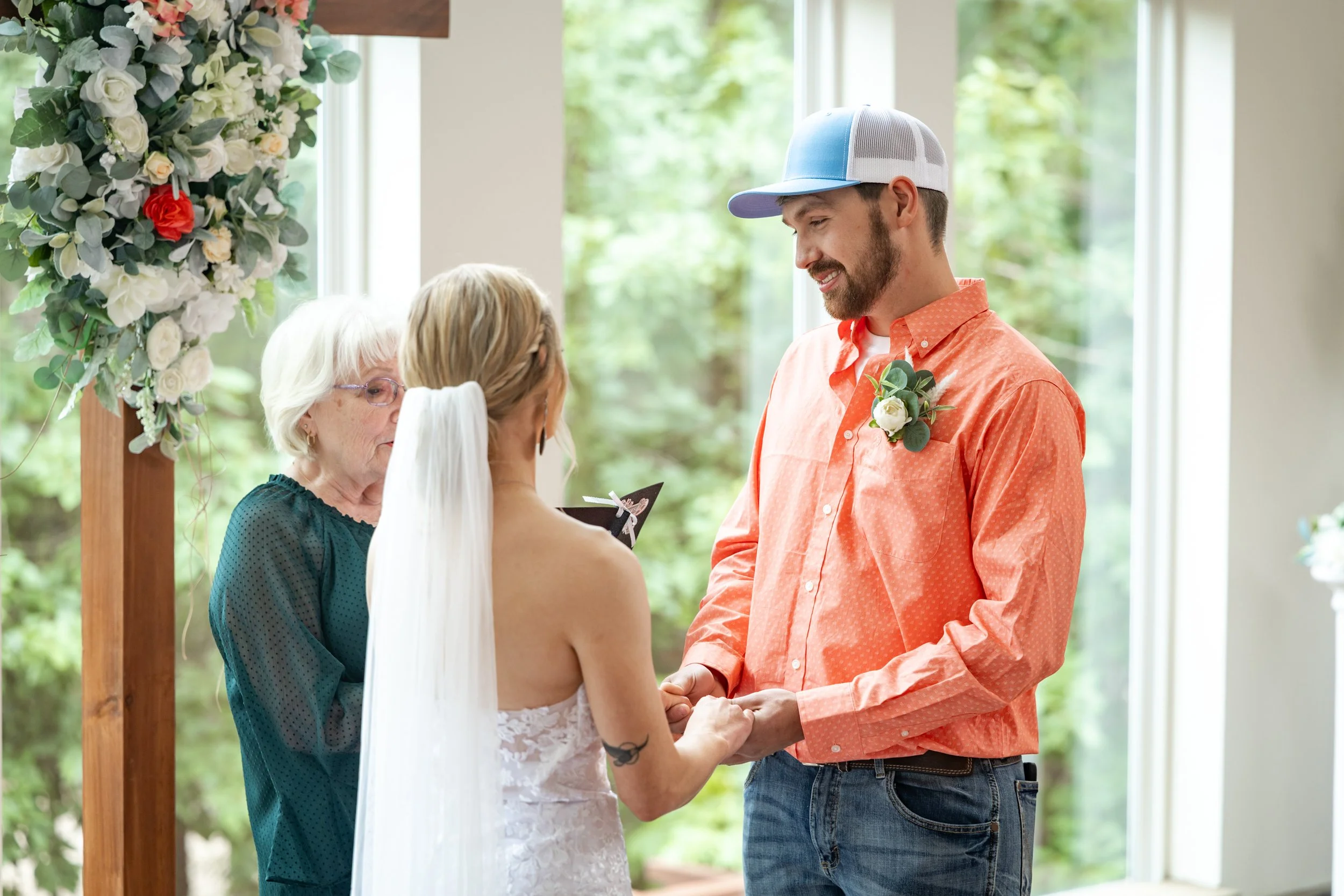 A man and woman exchange wedding vows outdoors, with an officiant standing nearby. The man wears a light blue and white cap, an orange shirt with a boutonniere, and jeans. The woman wears a white lace dress and has a tattoo on her arm. They are holdi