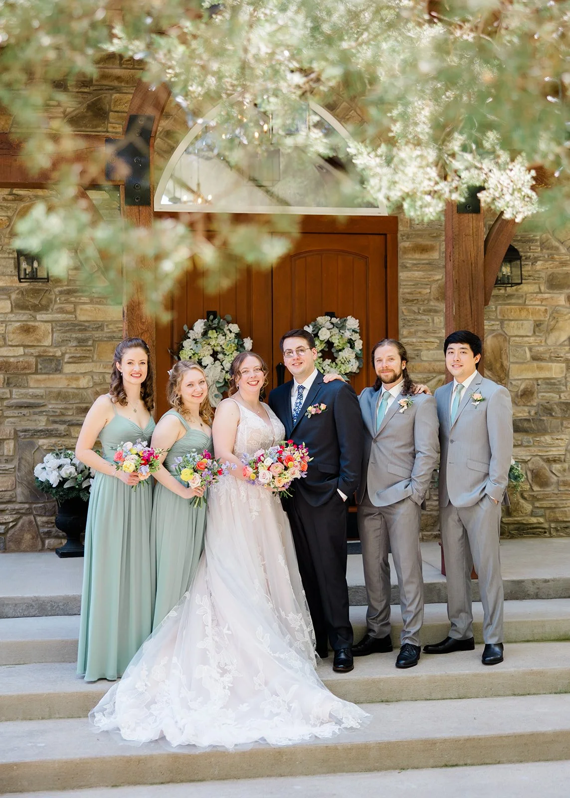 A wedding party standing on steps outside a stone building with a wooden door and floral wreaths, with blurred tree branches in the foreground.