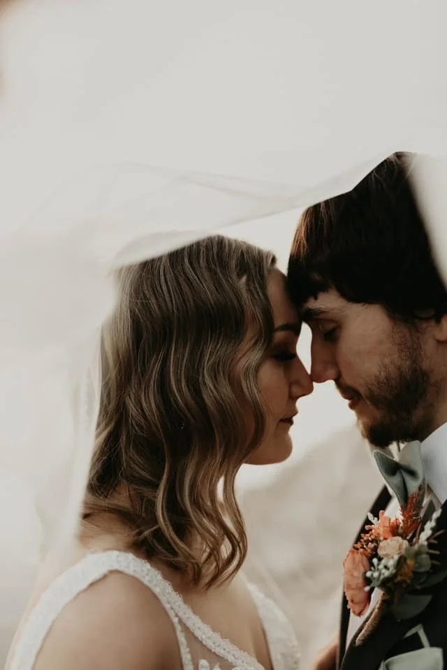 Close-up of a bride and groom with their foreheads touching, eyes closed, under a white veil during their wedding ceremony.