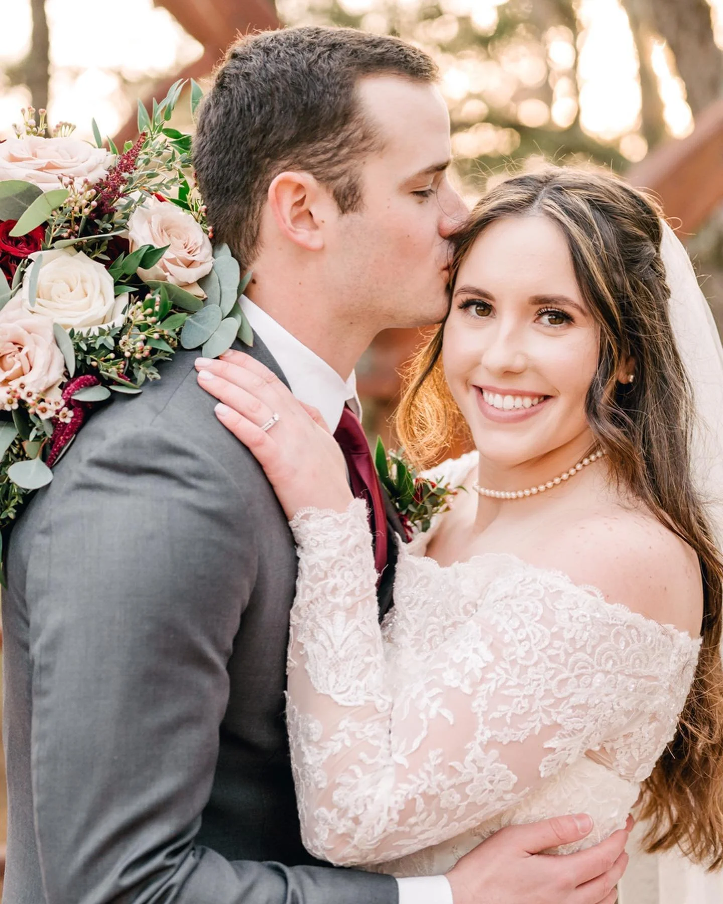 A newlywed couple during their wedding, with the groom kissing the bride on the forehead, the bride smiling at the camera, holding a bouquet of flowers, outdoors with sunlight filtering through trees.