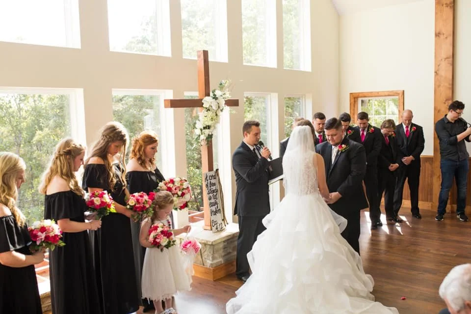 A wedding ceremony inside a church with the bride and groom exchanging vows, surrounded by bridesmaids and groomsmen, with a large wooden cross decorated with flowers in the background.