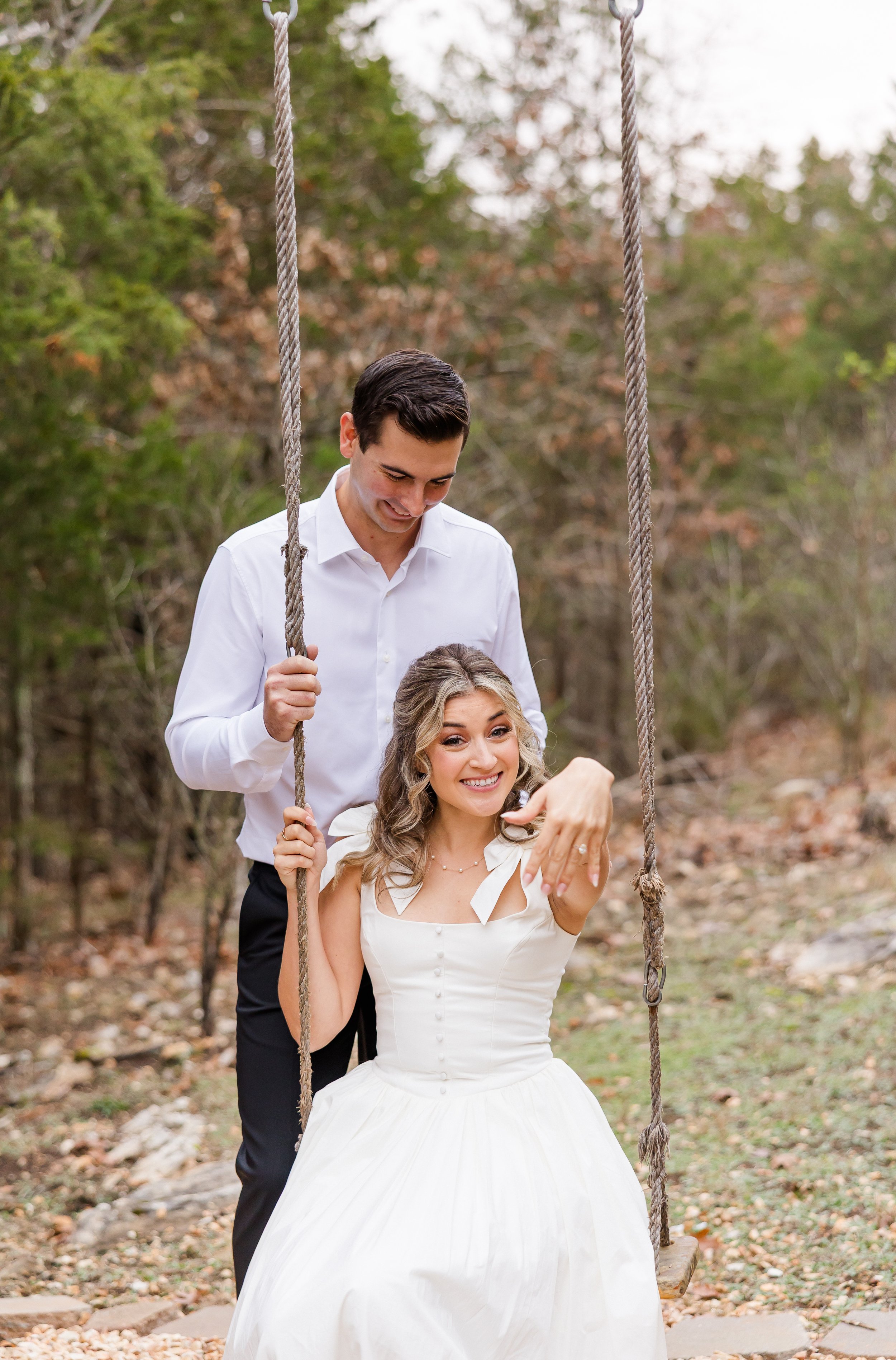 A happy woman in a white dress sitting on a swing in a wooded outdoor setting, with a man in a white shirt standing behind her holding the swing ropes.