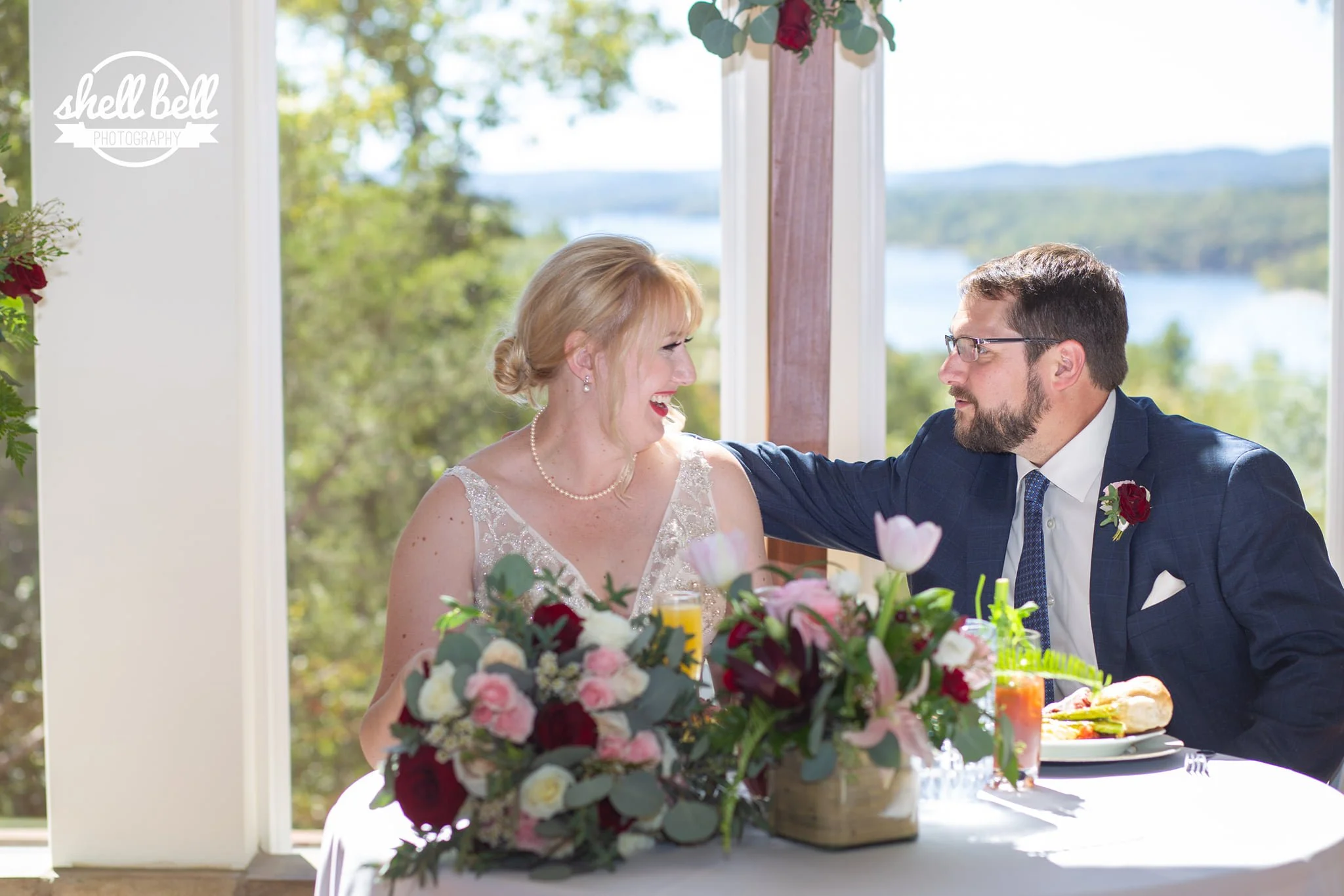 A woman and a man at a wedding reception, sitting at a table with a large floral centerpiece, food, and beverages, with a scenic outdoor view in the background.