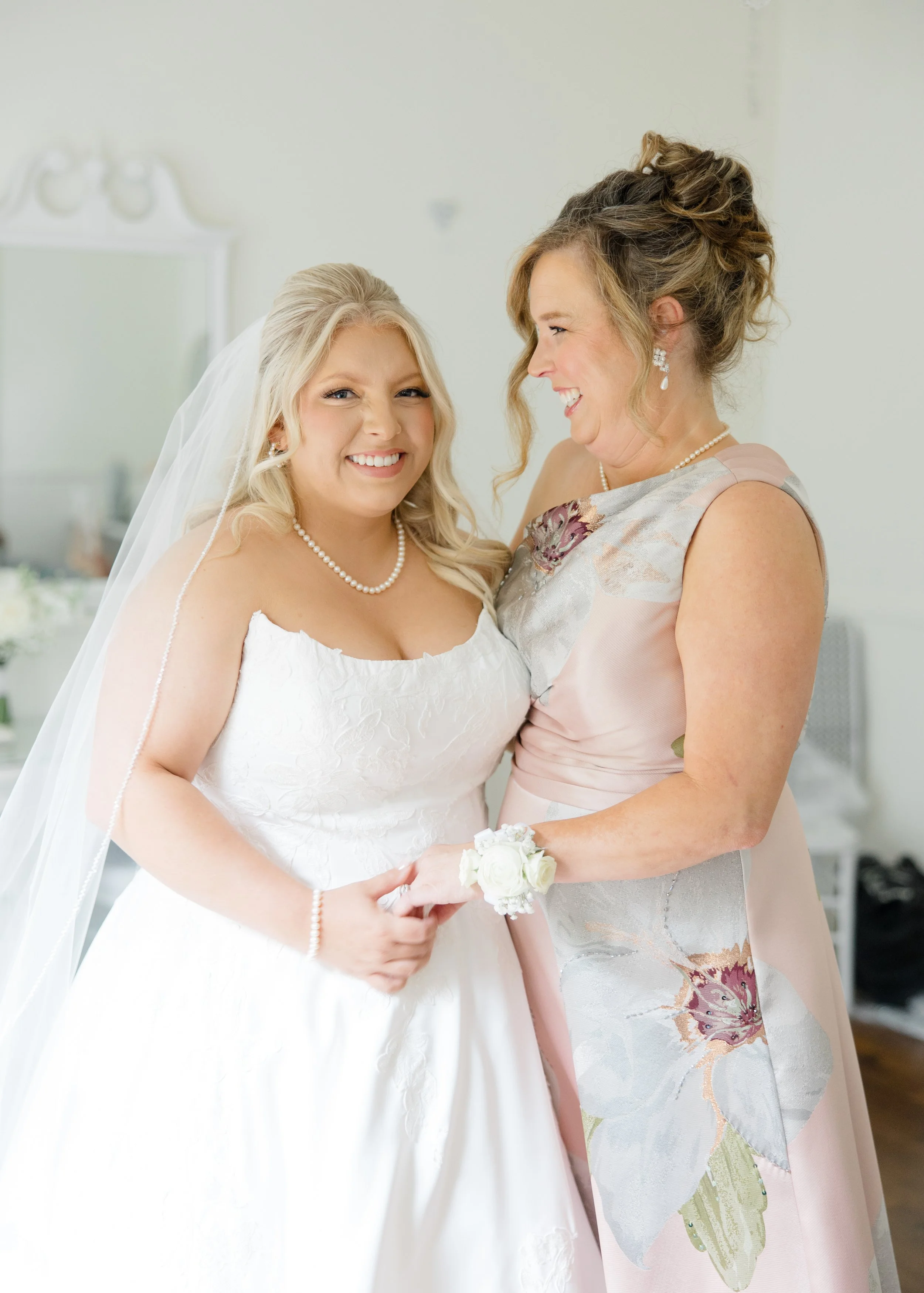 A bride with blonde hair wearing a white wedding dress and pearl jewelry, holding a small bouquet of white roses, smiling at a woman in a floral dress with butterfly pattern and pearl jewelry, both smiling and looking at each other.