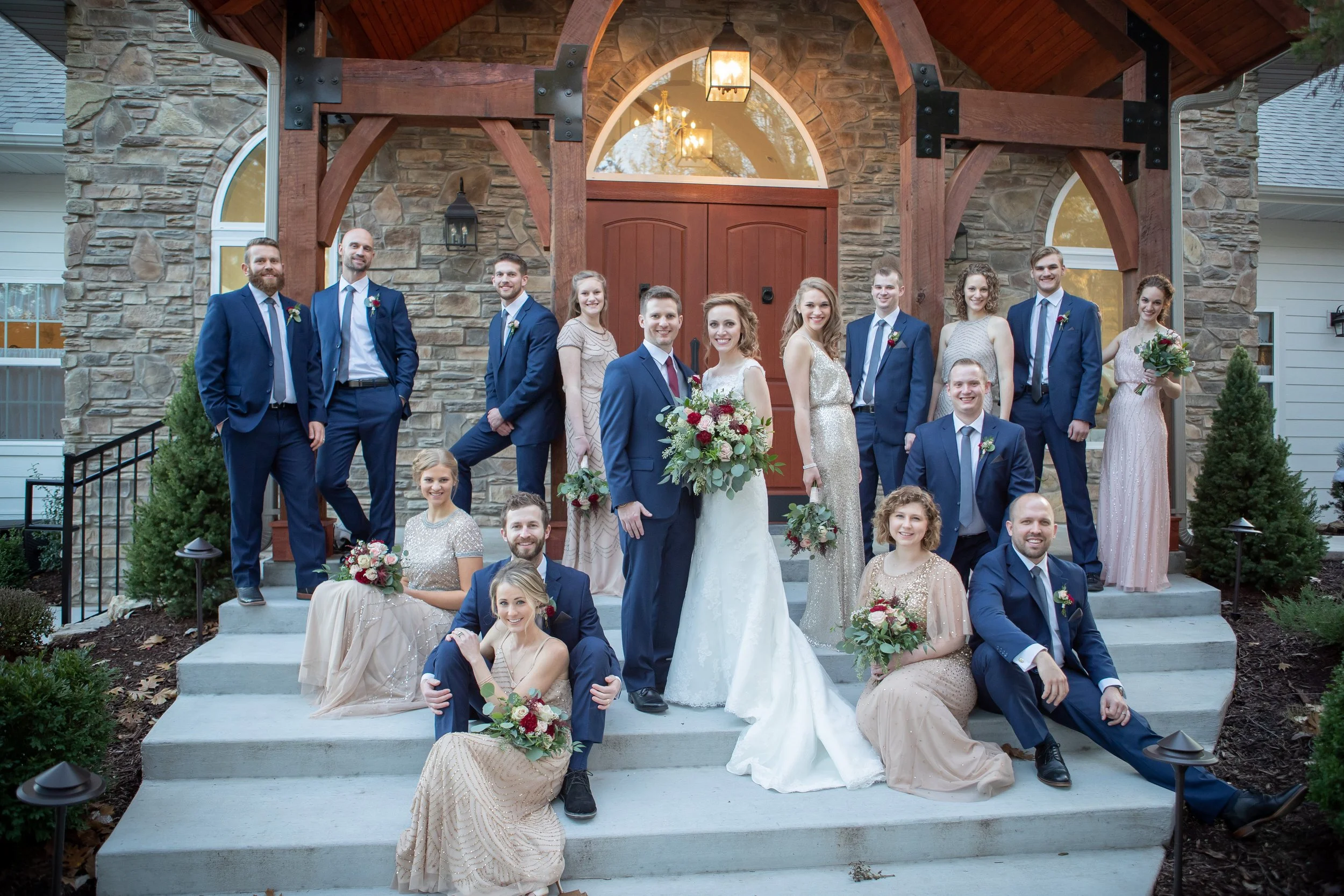 A large group of people dressed in formal attire, including a bride in a white wedding dress holding a bouquet of flowers, and a groom in a navy suit, standing on the steps of a stone building with wooden accents and large windows. The group appears 