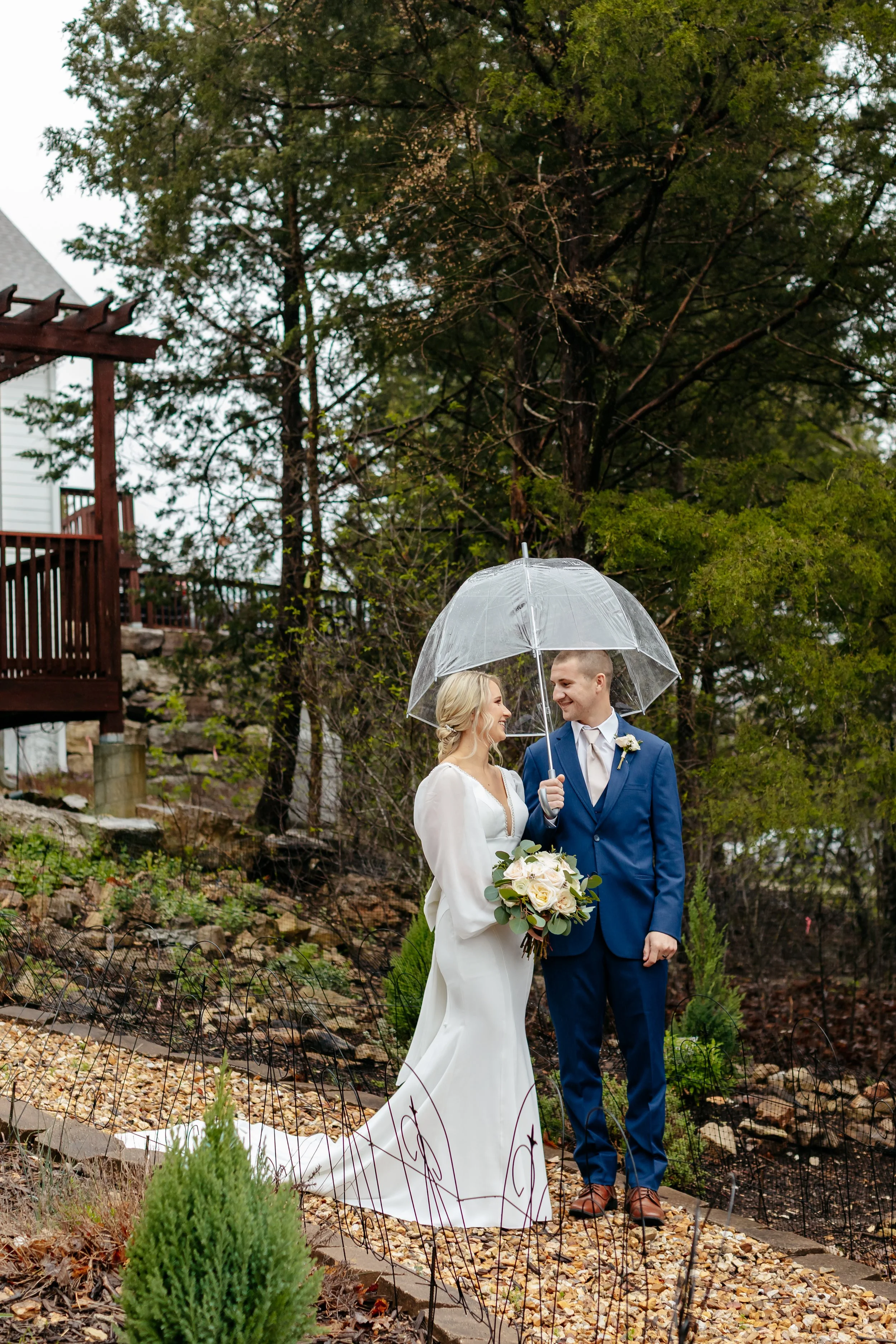 A bride and groom stand close together under an umbrella in a garden setting, smiling and gazing into each other's eyes, with trees and a house in the background.