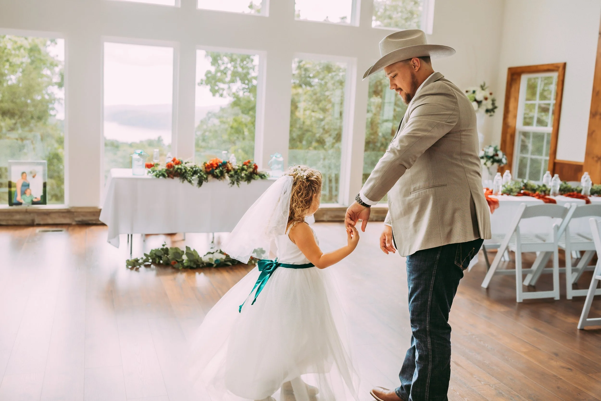 A man in a light gray suit and cowboy hat dancing with a young girl in a white dress with a teal sash at a wedding reception inside a bright room with large windows and wooden floors.