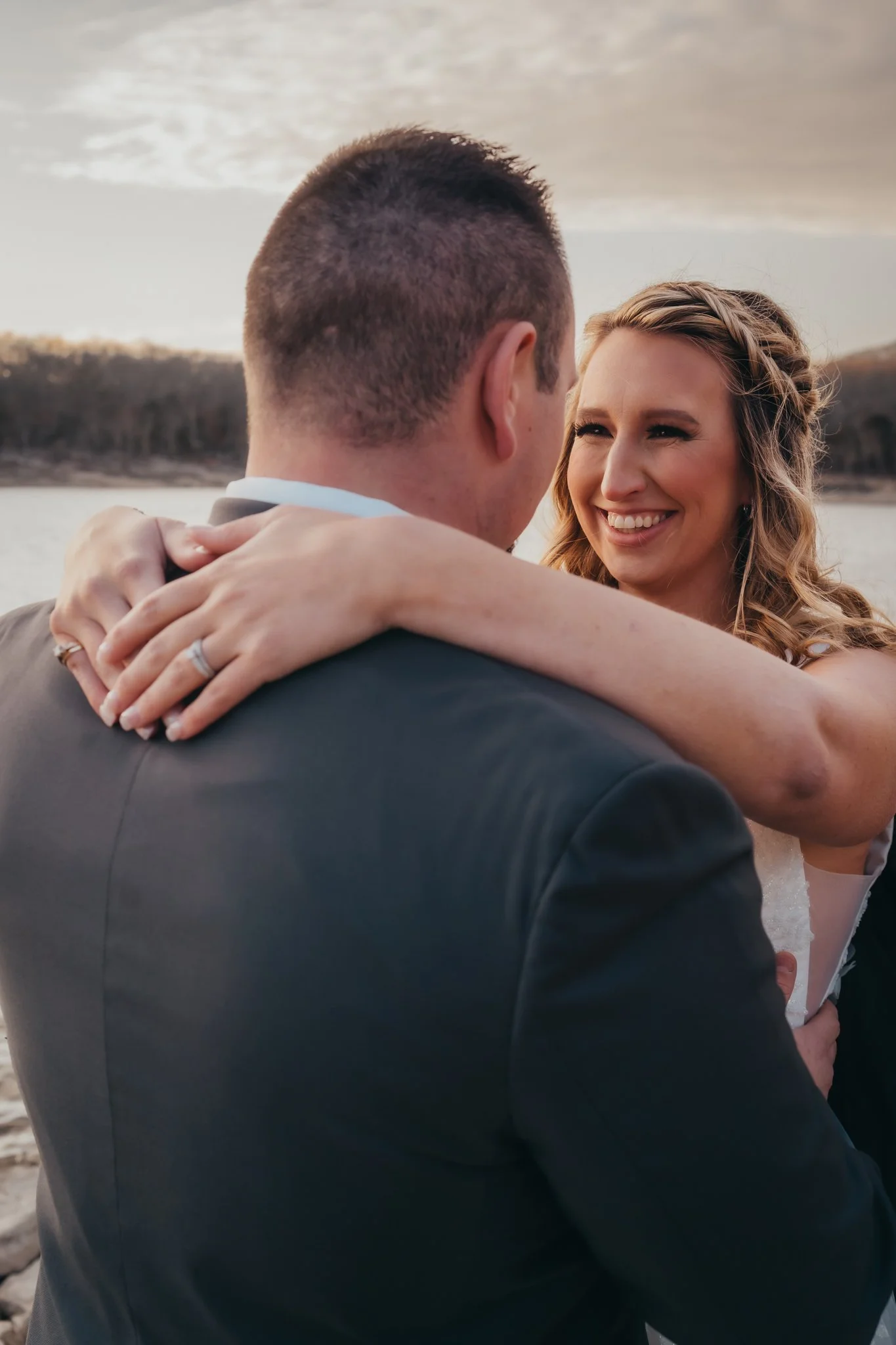 A couple embracing outdoors during sunset by a body of water, with trees and hills in the background.