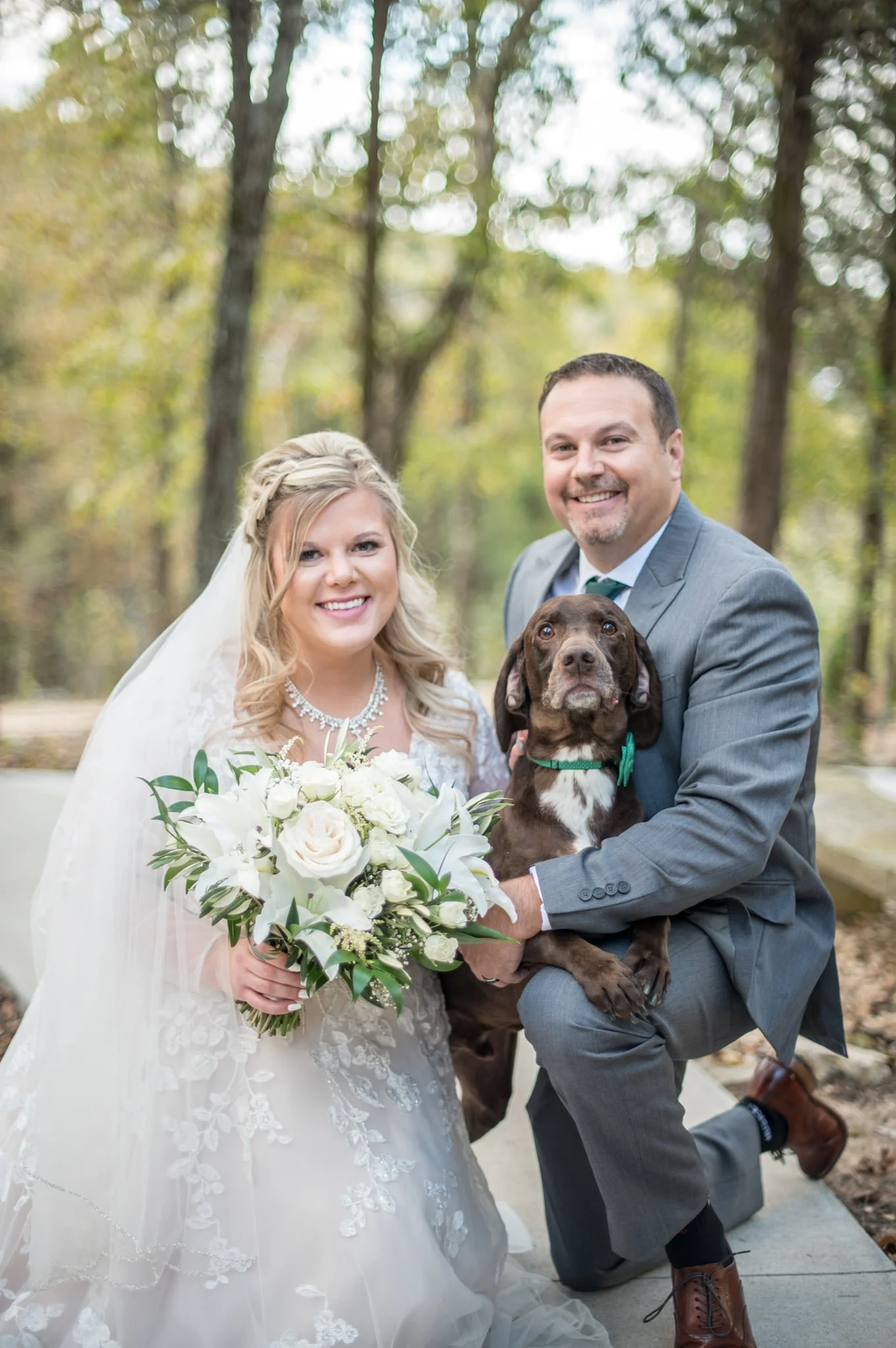 A bride and groom with a dog outdoors in a wooded area, with autumn leaves in the background, during a wedding celebration.