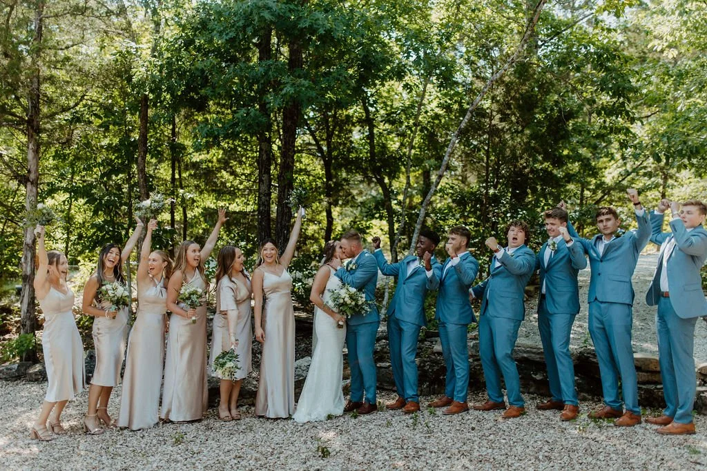 A wedding party standing outdoors in a wooded area, with the bride and groom kissing in the center. Bridesmaids and groomsmen are on each side, with the bridesmaids raising their flower bouquets and the groomsmen raising their fists in celebration.
