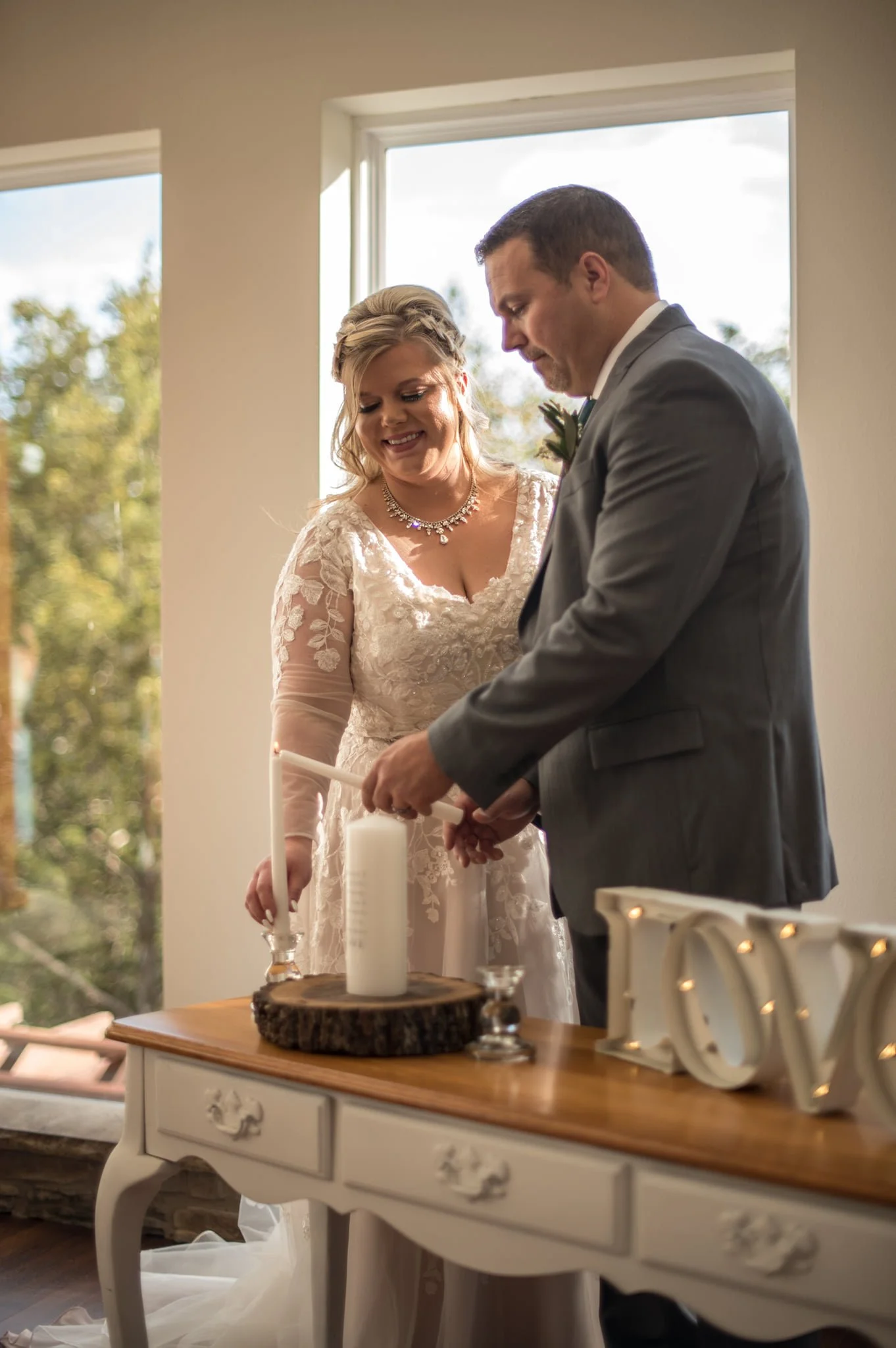 A bride and groom participating in a candle lighting ceremony during their wedding, standing in front of windows with natural light.